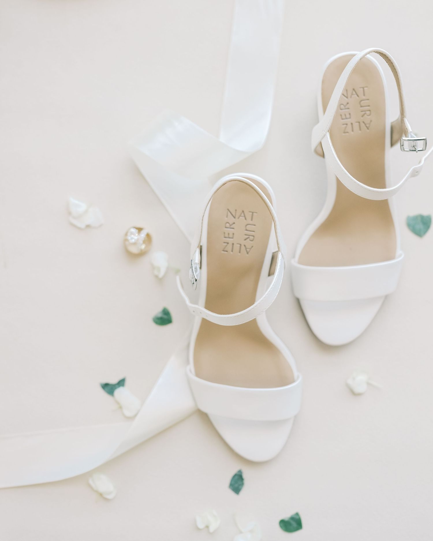 White bridal block heel sandals with ankle straps displayed on a neutral background with scattered flower petals.