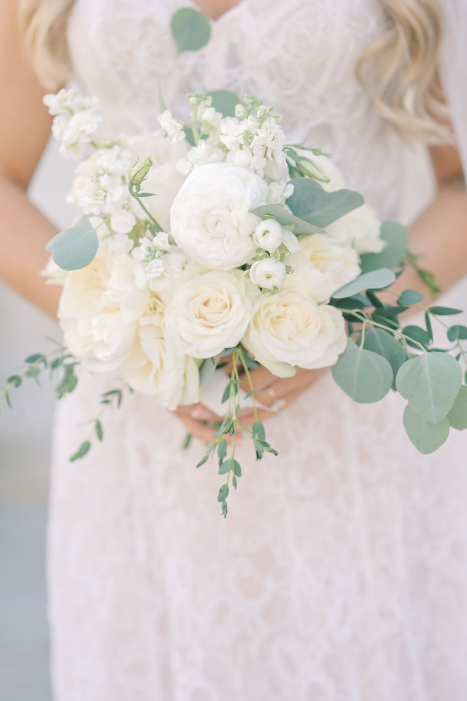Bridal bouquet of white roses and eucalyptus held by bride in lace dress.