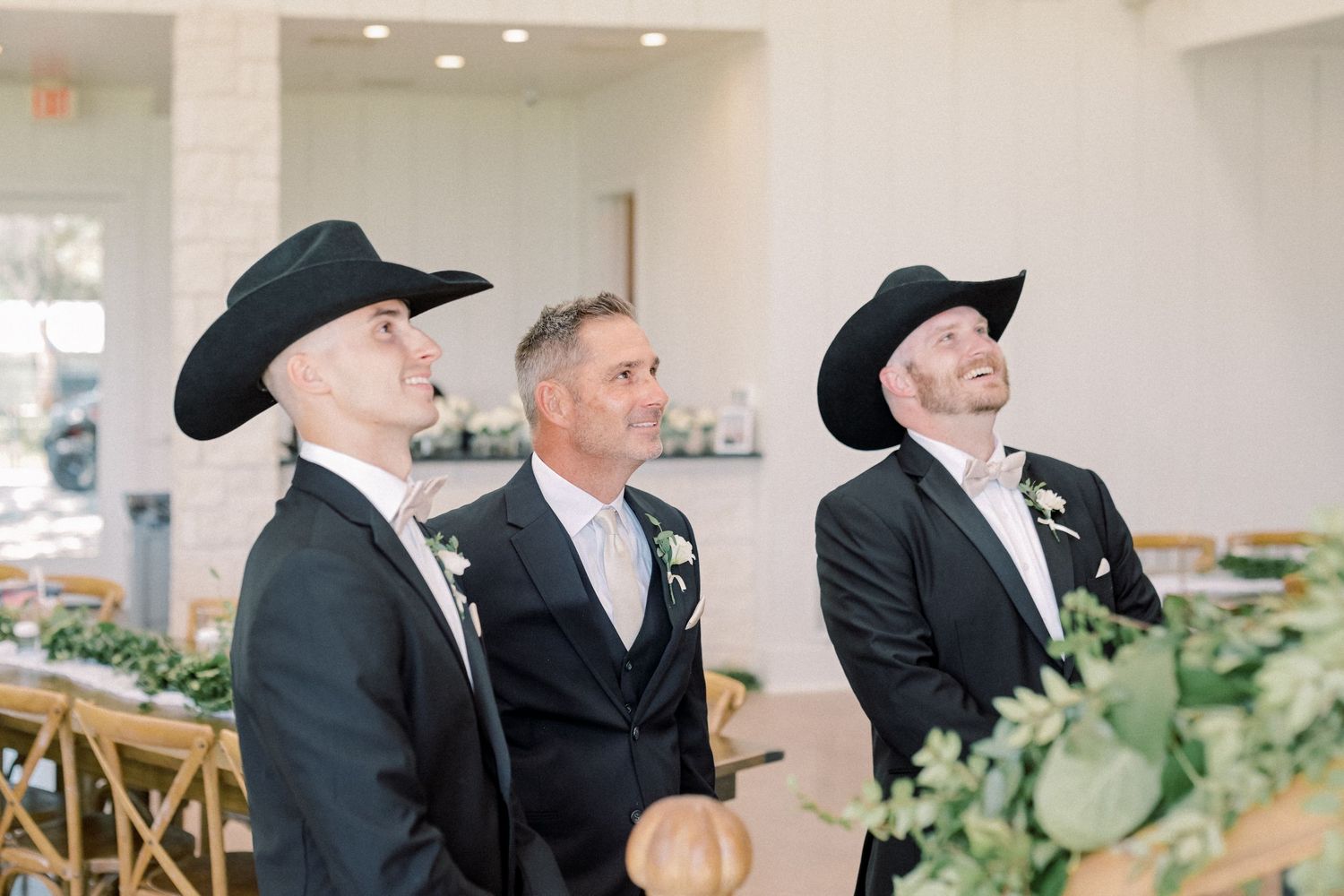 Three groomsmen in black suits and cowboy hats smile during an indoor wedding ceremony.
