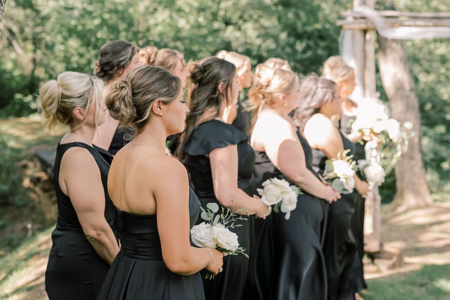 Bridesmaids in black dresses line up holding white bouquets at rustic outdoor ceremony.