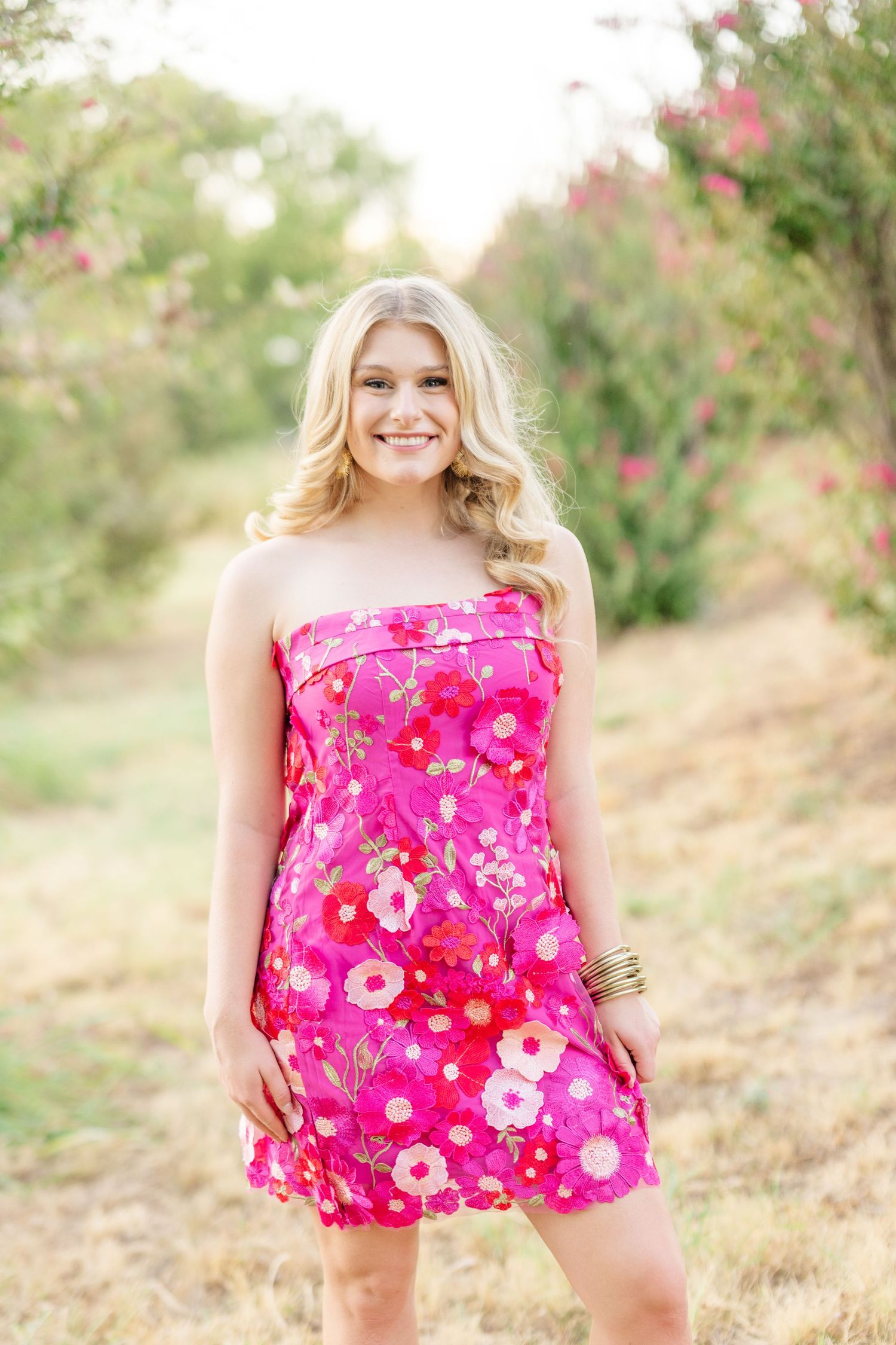 Smiling blonde woman in a pink floral strapless mini dress standing outdoors in a garden setting.