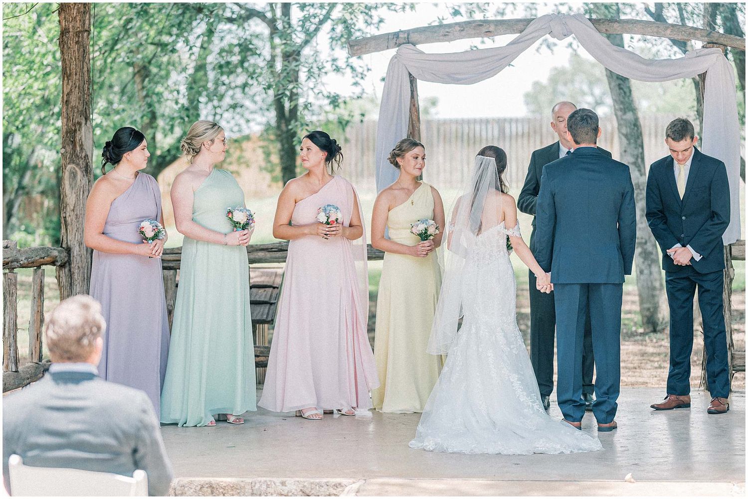 Wedding party in pastel dresses and navy suits pose outdoors under trees.