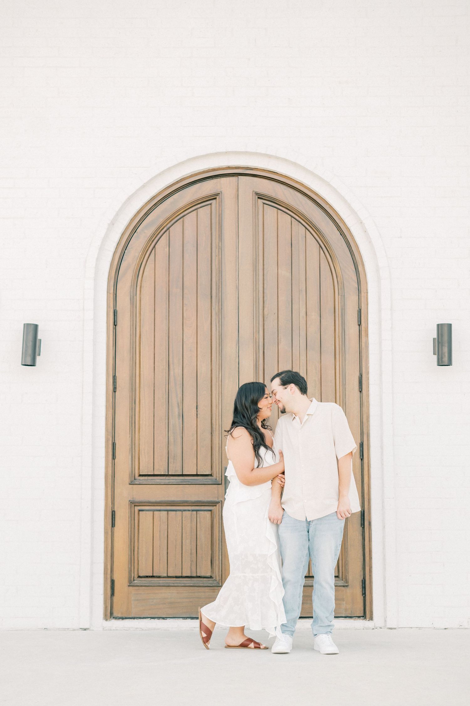A couple in light-colored outfits sharing an intimate moment against a white wall with wooden arched doors.
