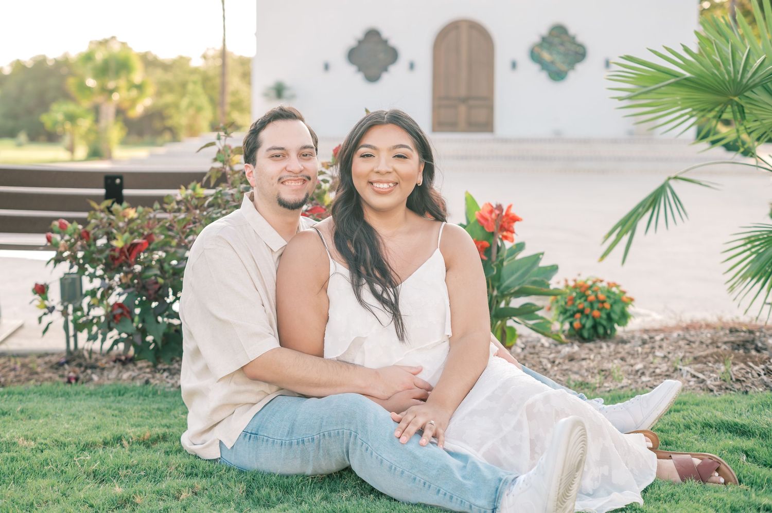 A couple in white outfits shares joyful moments together during an outdoor photo session in a garden setting.