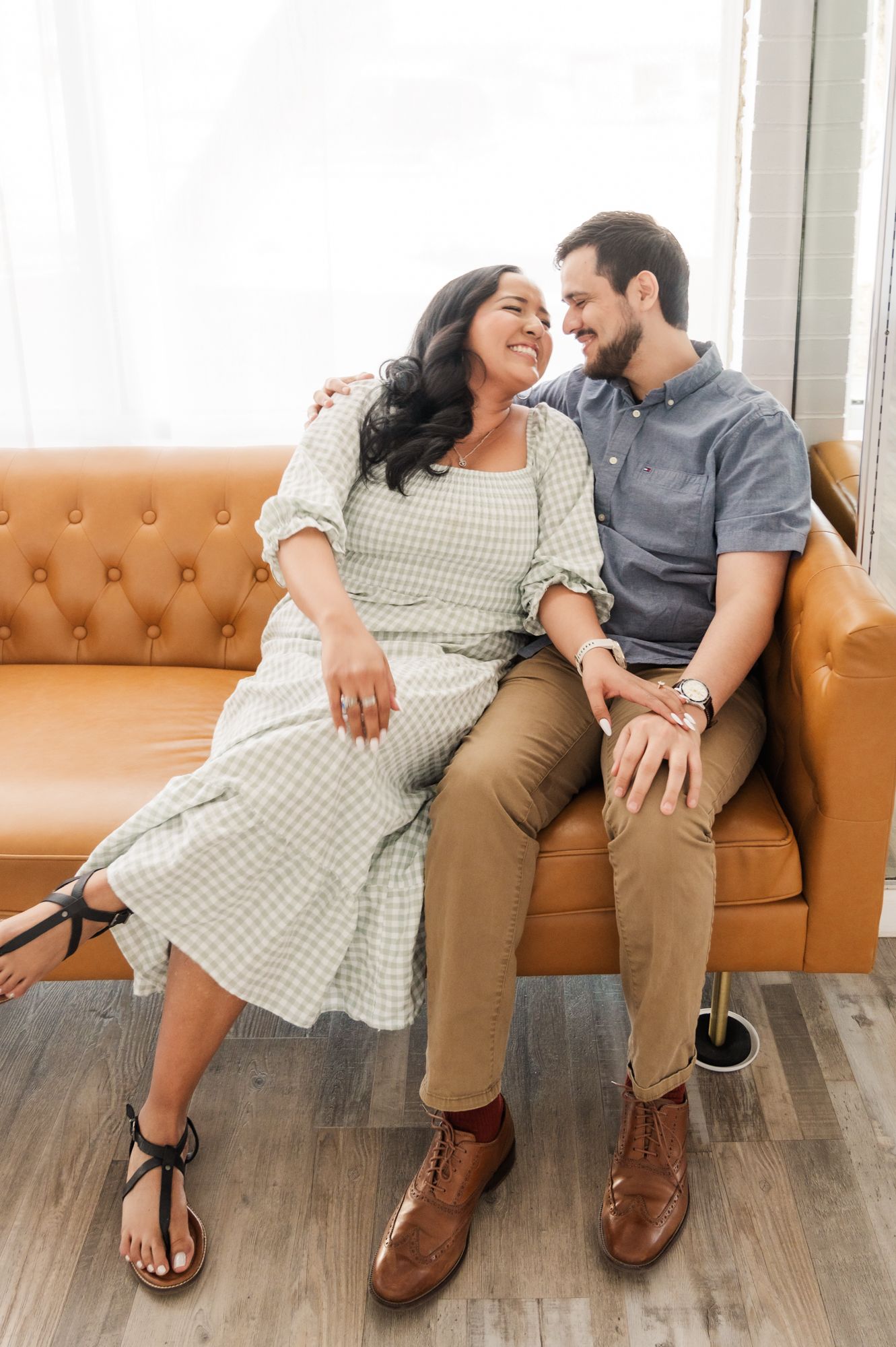 A joyful couple laughs together while relaxing on a tufted leather sofa in a sunlit room.