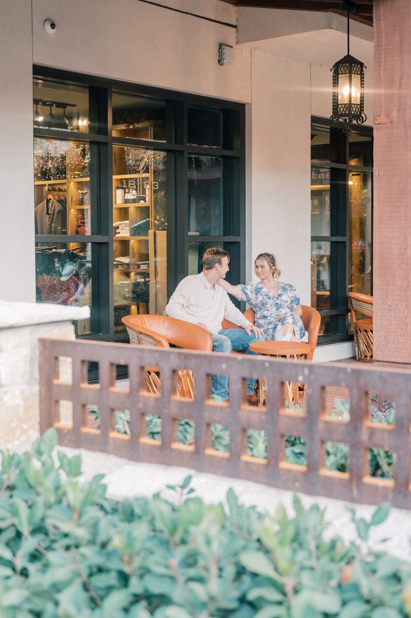 A couple sits together on an outdoor patio with orange chairs and greenery in the foreground.
