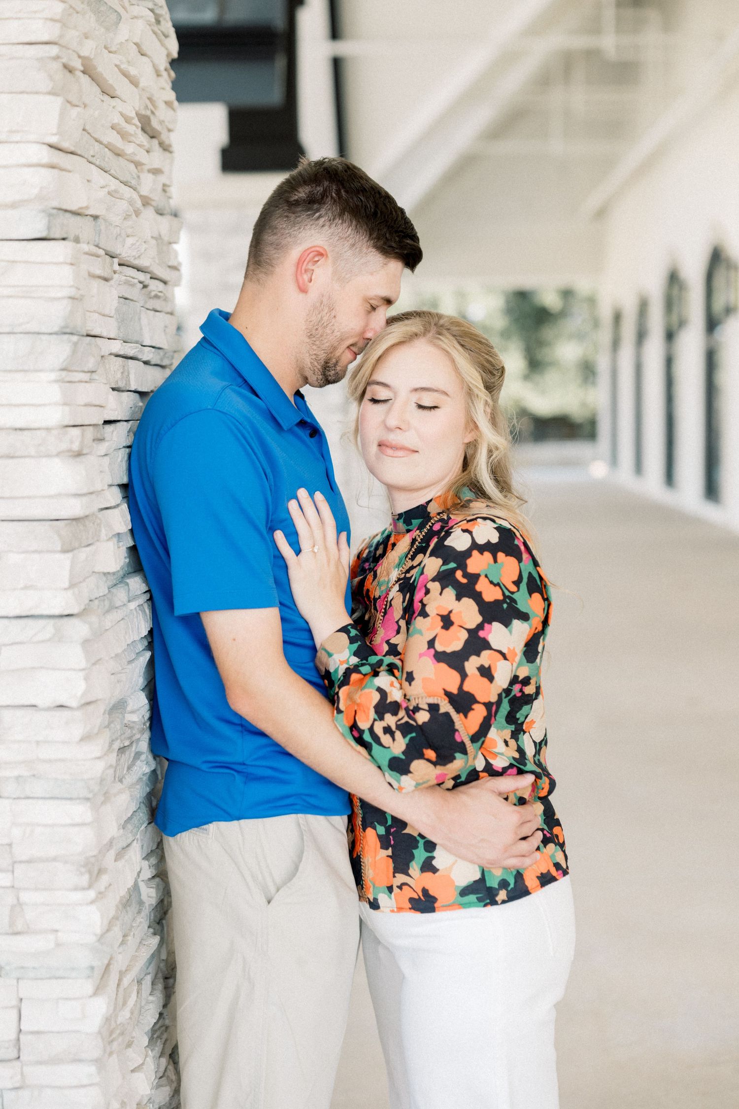 Romantic couple leaning against stone column, man nuzzling woman in colorful floral blouse and white pants.