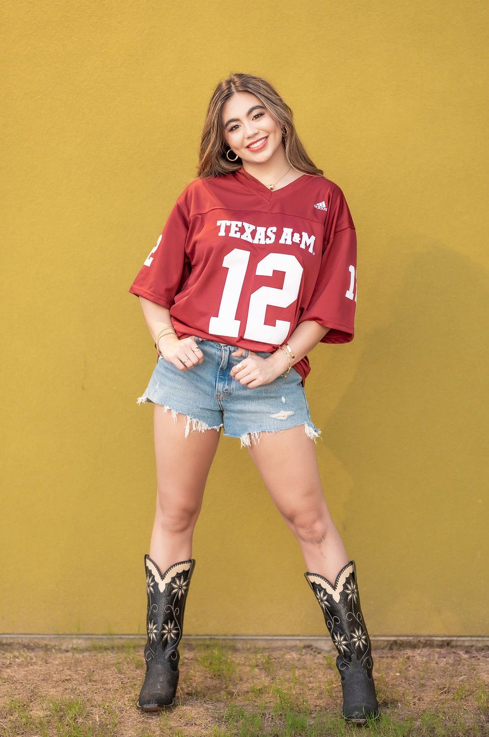 Smiling young woman wearing a Texas A&M #12 jersey, denim shorts, and cowboy boots against a yellow wall.