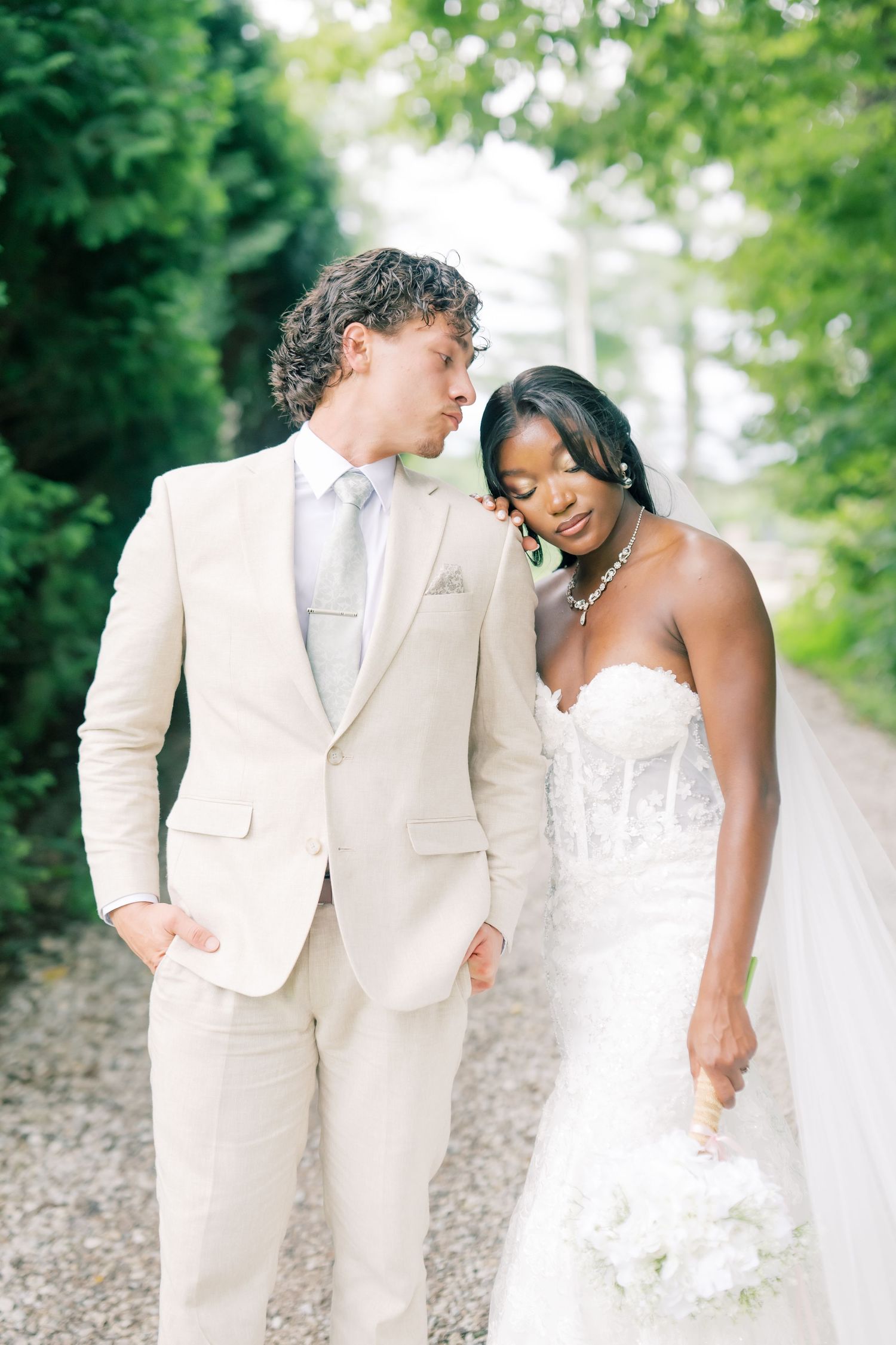 Interracial wedding couple in romantic pose, groom in cream suit leaning toward bride in strapless lace gown with veil.