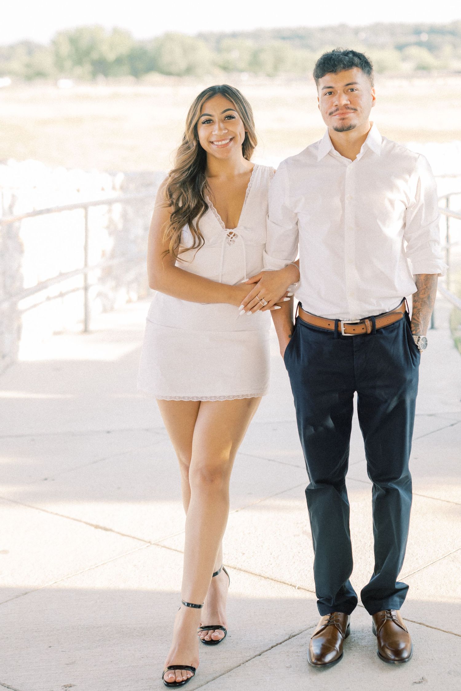 Couple posing outdoors in white dress and white shirt with navy pants for engagement photos.