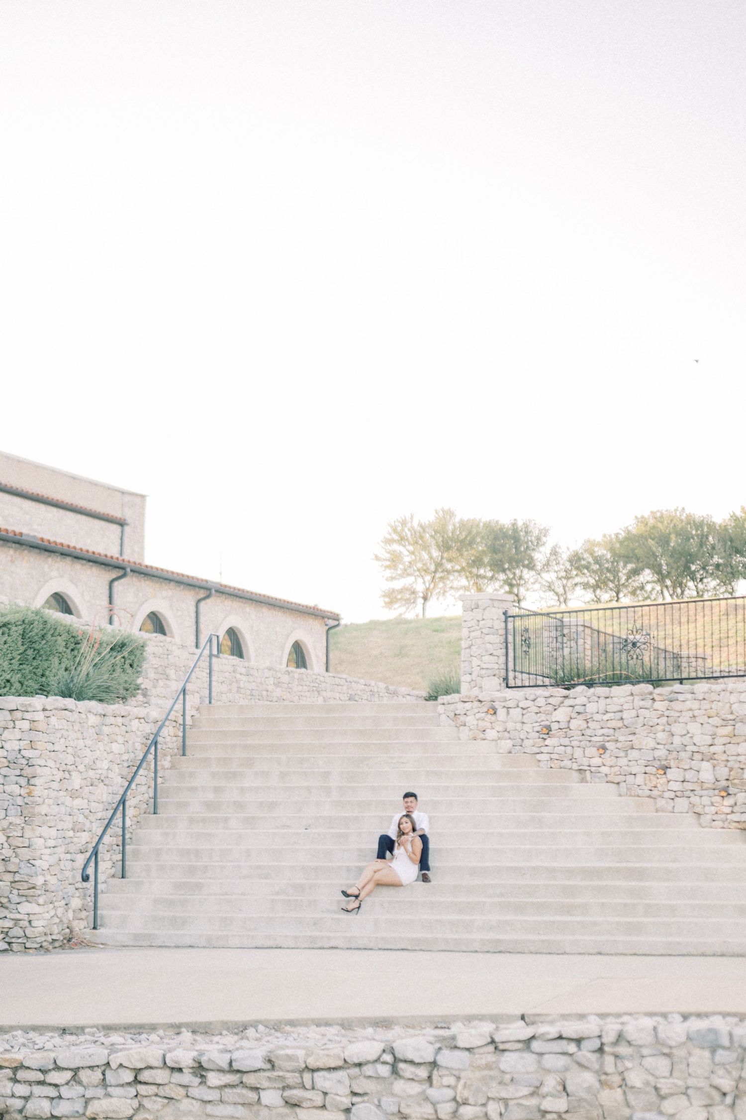 An engaged couple sits closely together on wide stone steps outside a rustic venue at golden hour.