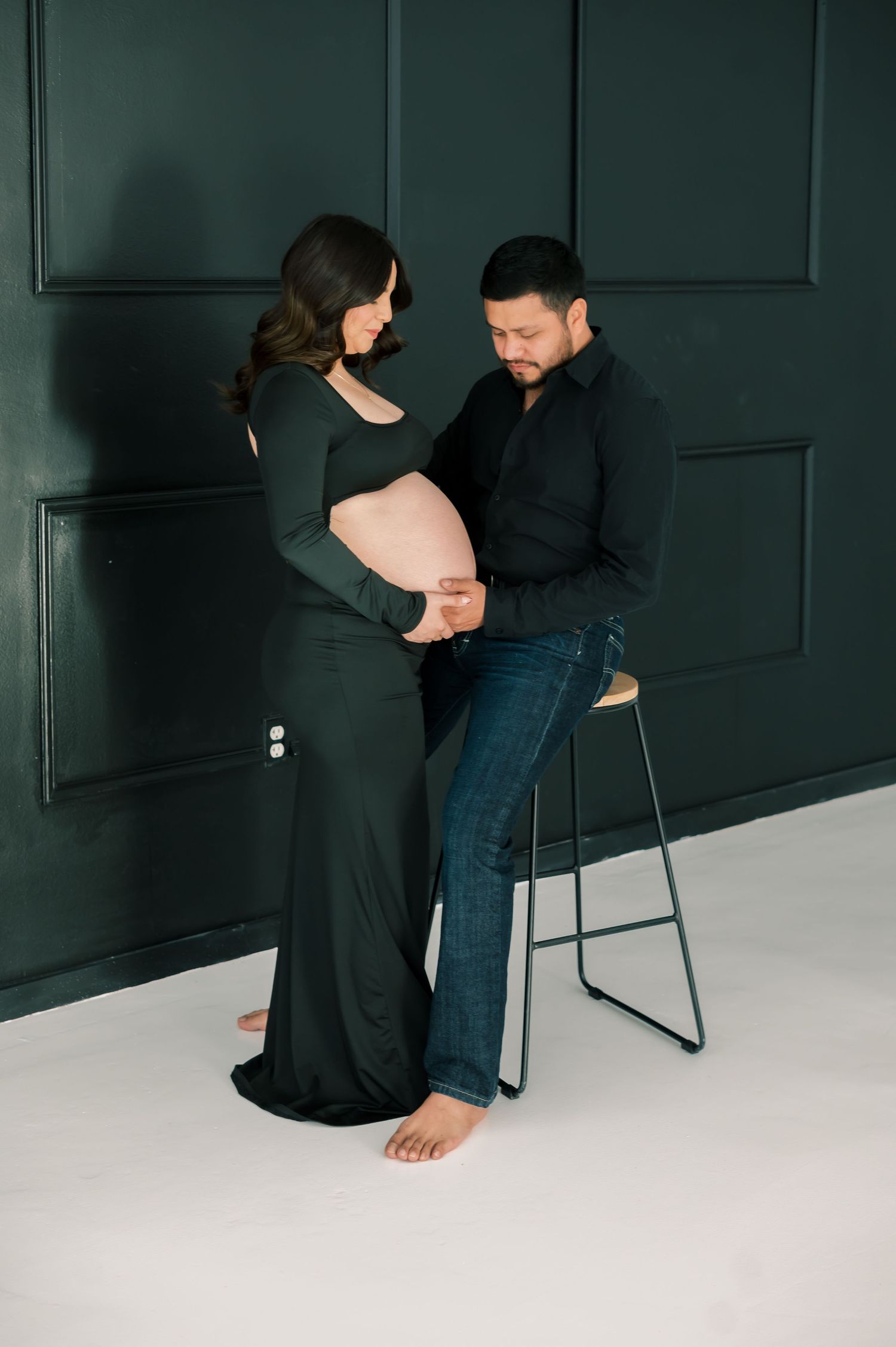Expecting couple in matching dark green and black outfits tenderly cradling pregnant belly during maternity photo session.