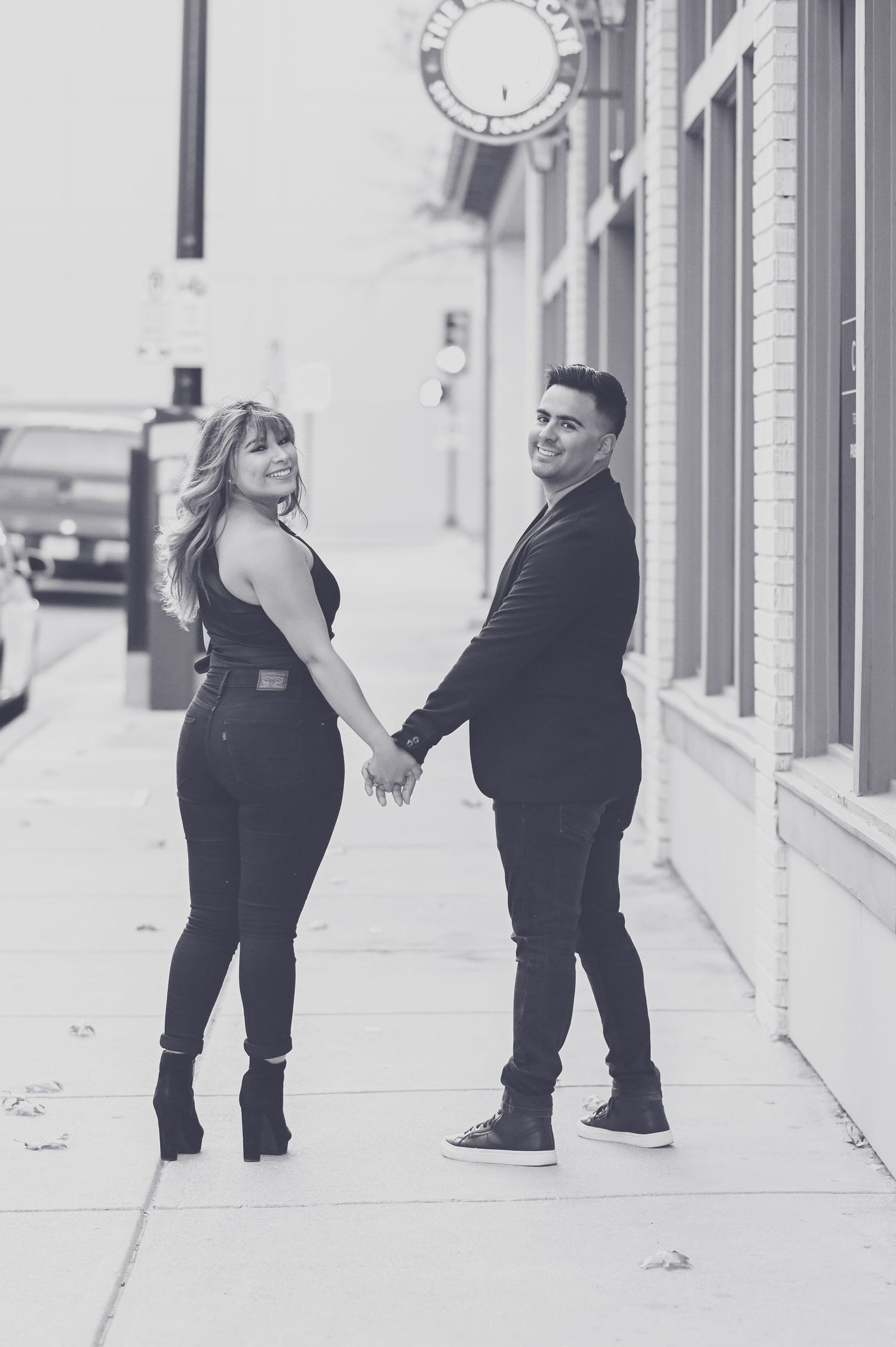 Black and white photo of a couple holding hands and looking back while walking on a sidewalk.