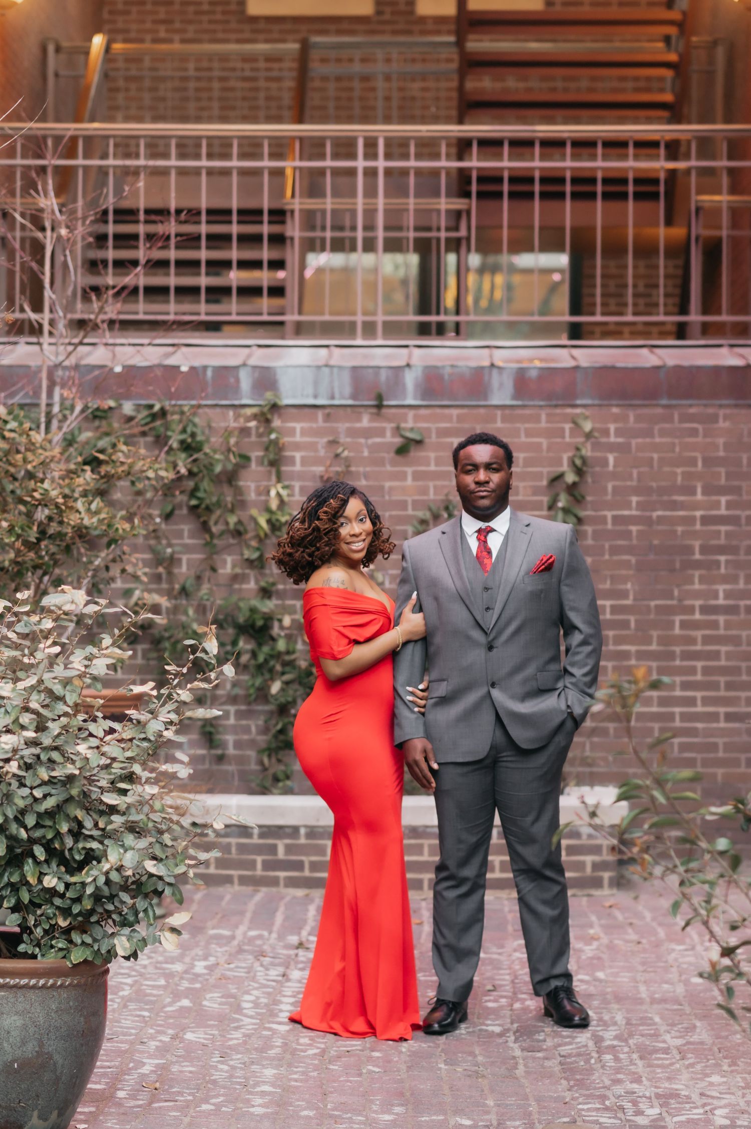 Elegant couple posing outdoors, woman in red off-shoulder gown and man in gray suit with red tie.