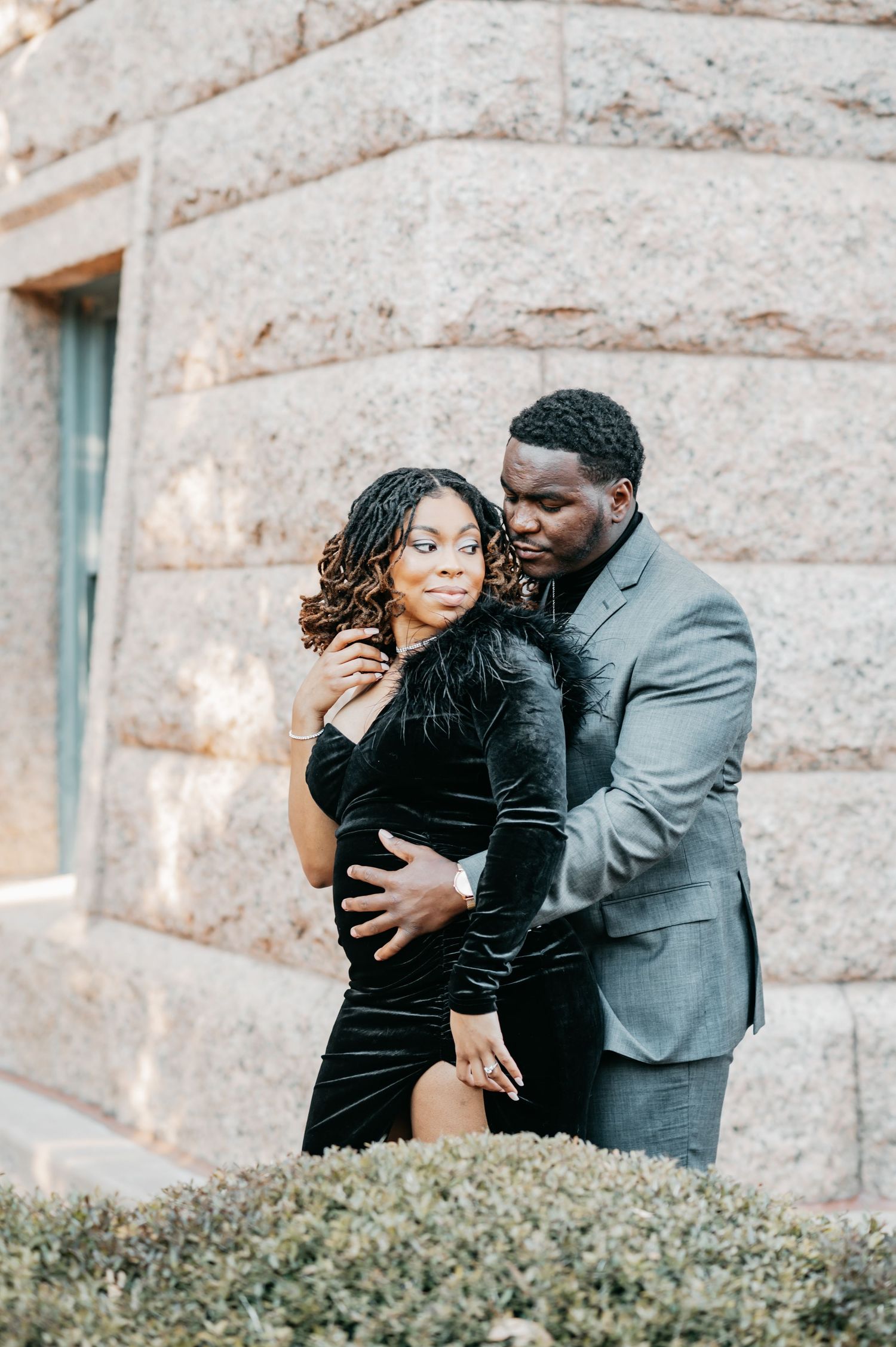 Couple posing elegantly outdoors, woman in black velvet dress, man in gray suit, against stone building.