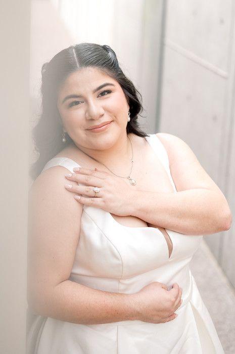 Elegant bride in white gown with diamond ring and necklace posing against light wall.