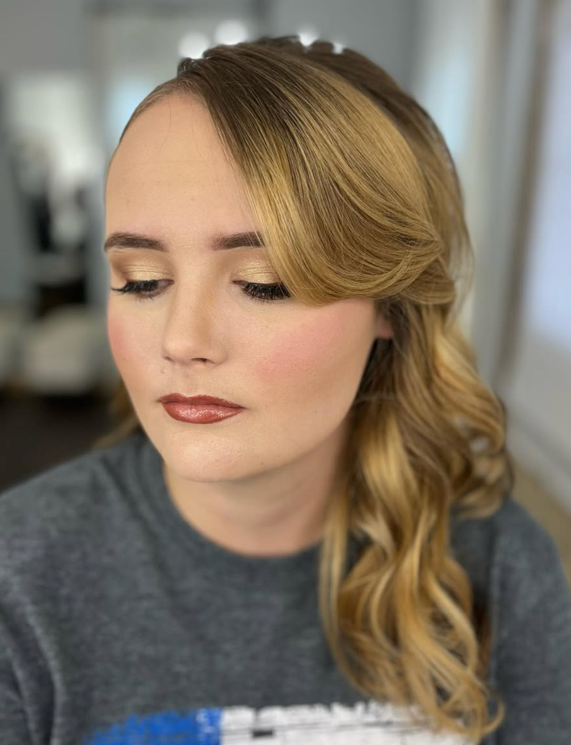 Woman with elegant side-swept curls and professional makeup featuring rosy blush and nude lips.