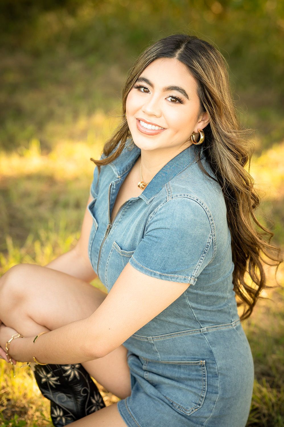 Young woman in denim dress smiling outdoors, sitting in sunlit green field with long wavy hair and gold jewelry.