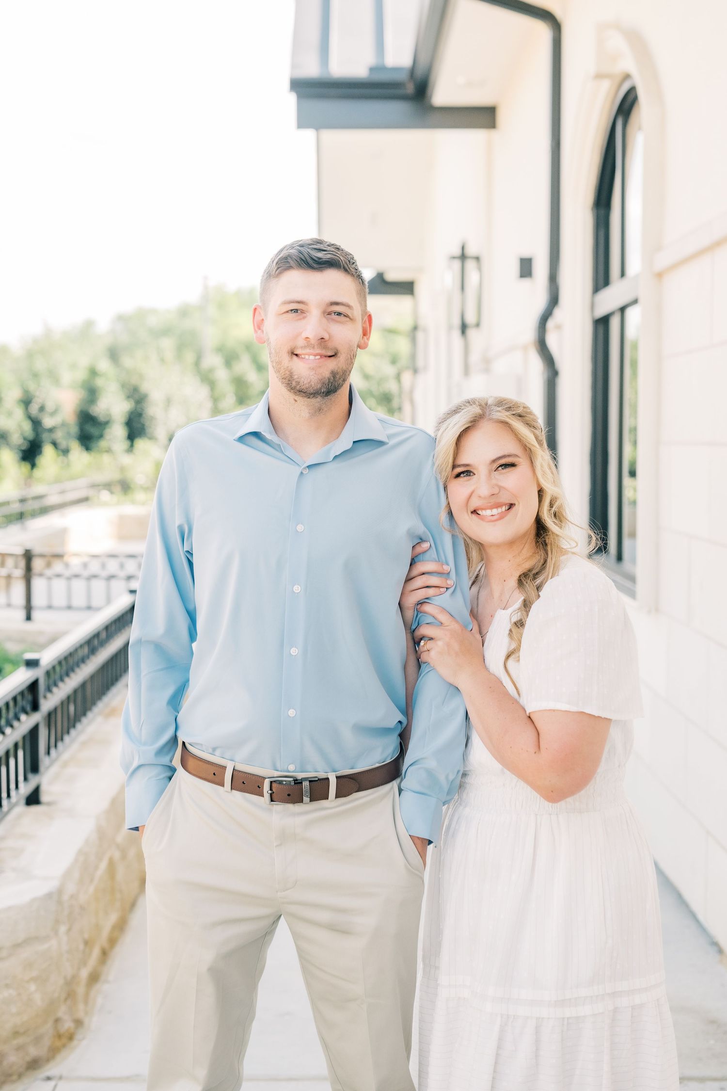 Smiling couple posing on a sunlit balcony, man in light blue shirt and woman in white dress.