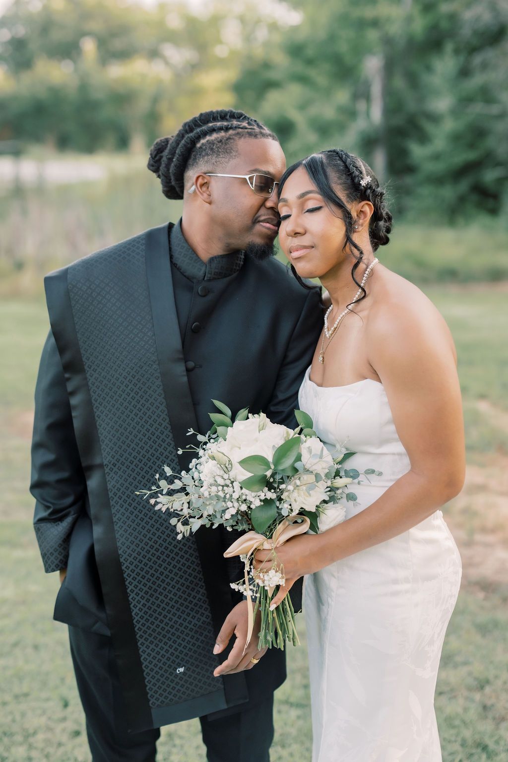 A Black bride and groom pose outdoors, she holds a white bouquet, he wears a black suit, both sharing an intimate moment.