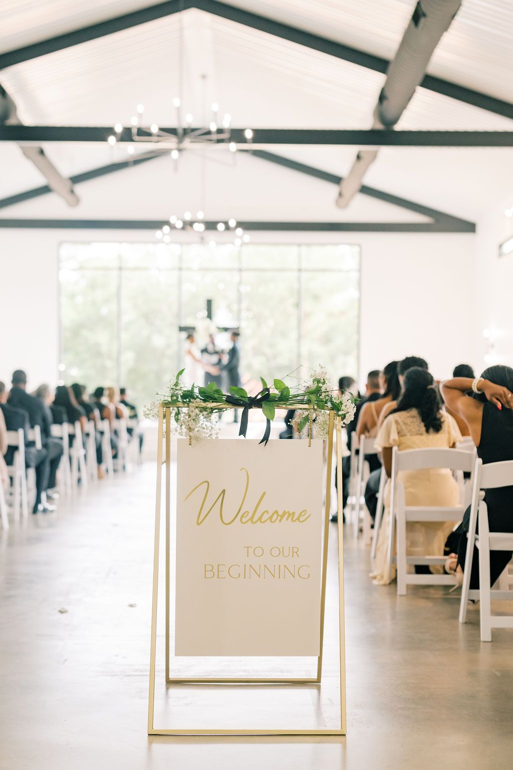Elegant wedding ceremony venue with a gold Welcome to Our Beginning sign adorned with greenery, white chairs, and vaulted ceiling.