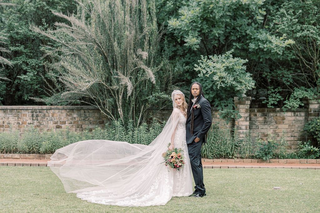 Bride in lace gown with dramatic flowing veil stands with groom in dark suit in garden with brick wall backdrop.
