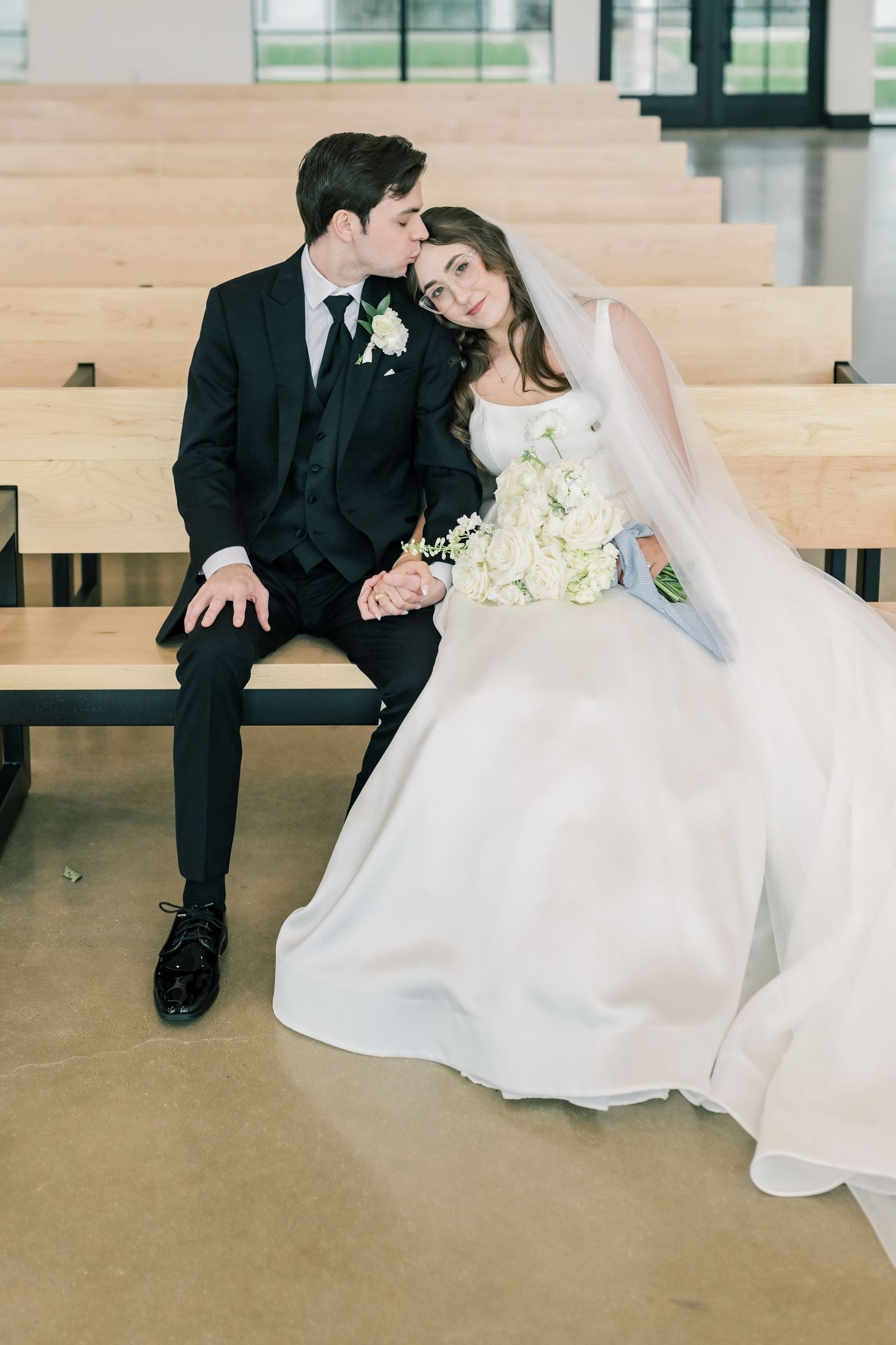 Bride and groom sit together on wooden pew holding hands with white floral bouquet.