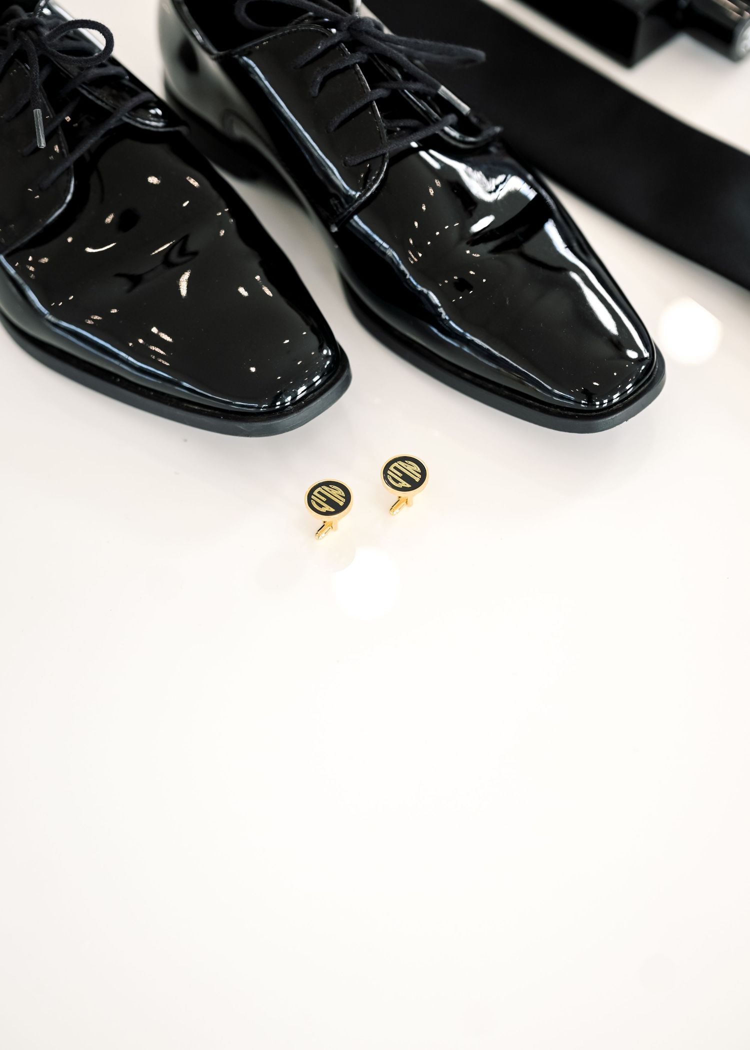 Black patent leather oxford dress shoes with gold monogram cufflinks on white surface.