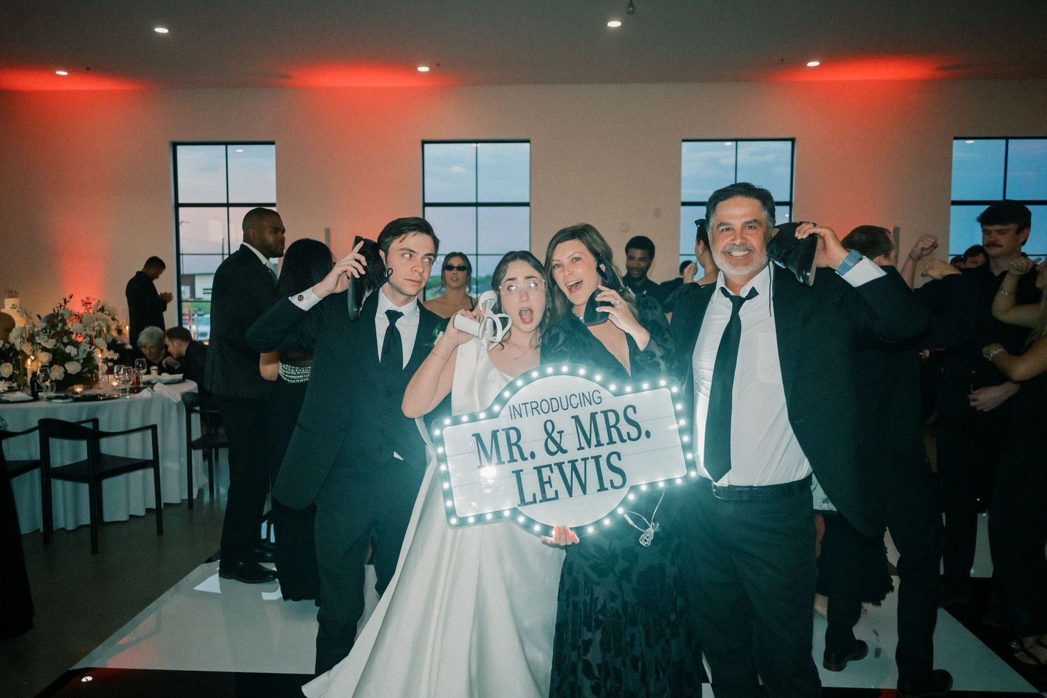 Newlyweds Mr. and Mrs. Lewis celebrate with friends holding illuminated sign at wedding reception.
