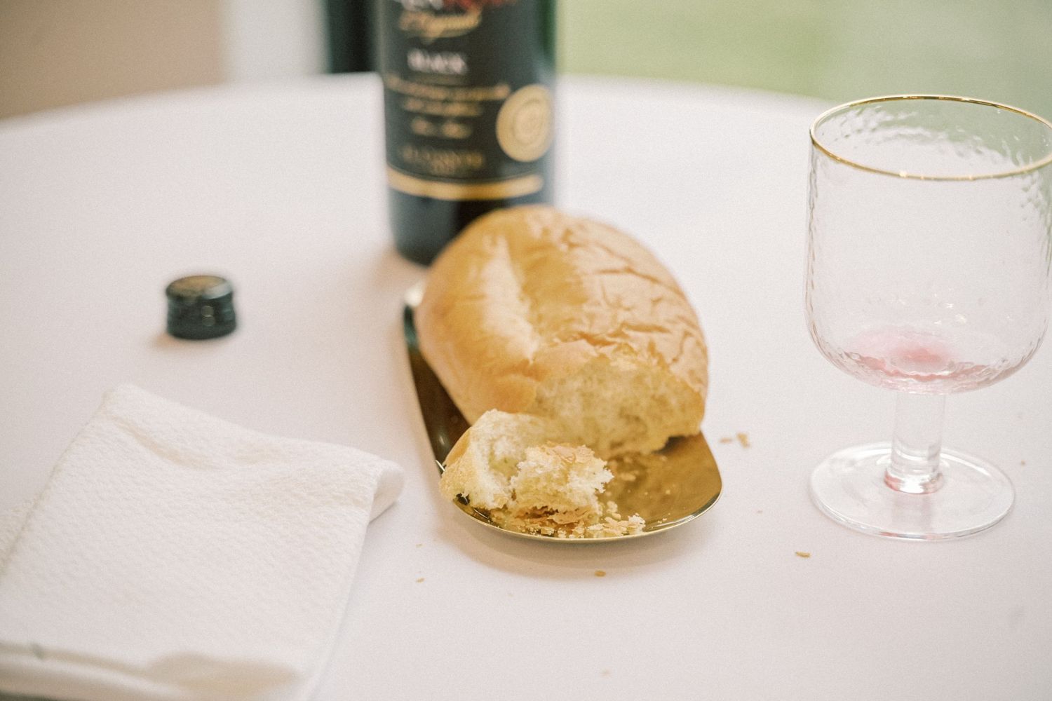 A bread roll with butter on a gold plate beside a wine glass and olive oil bottle on a white tablecloth.