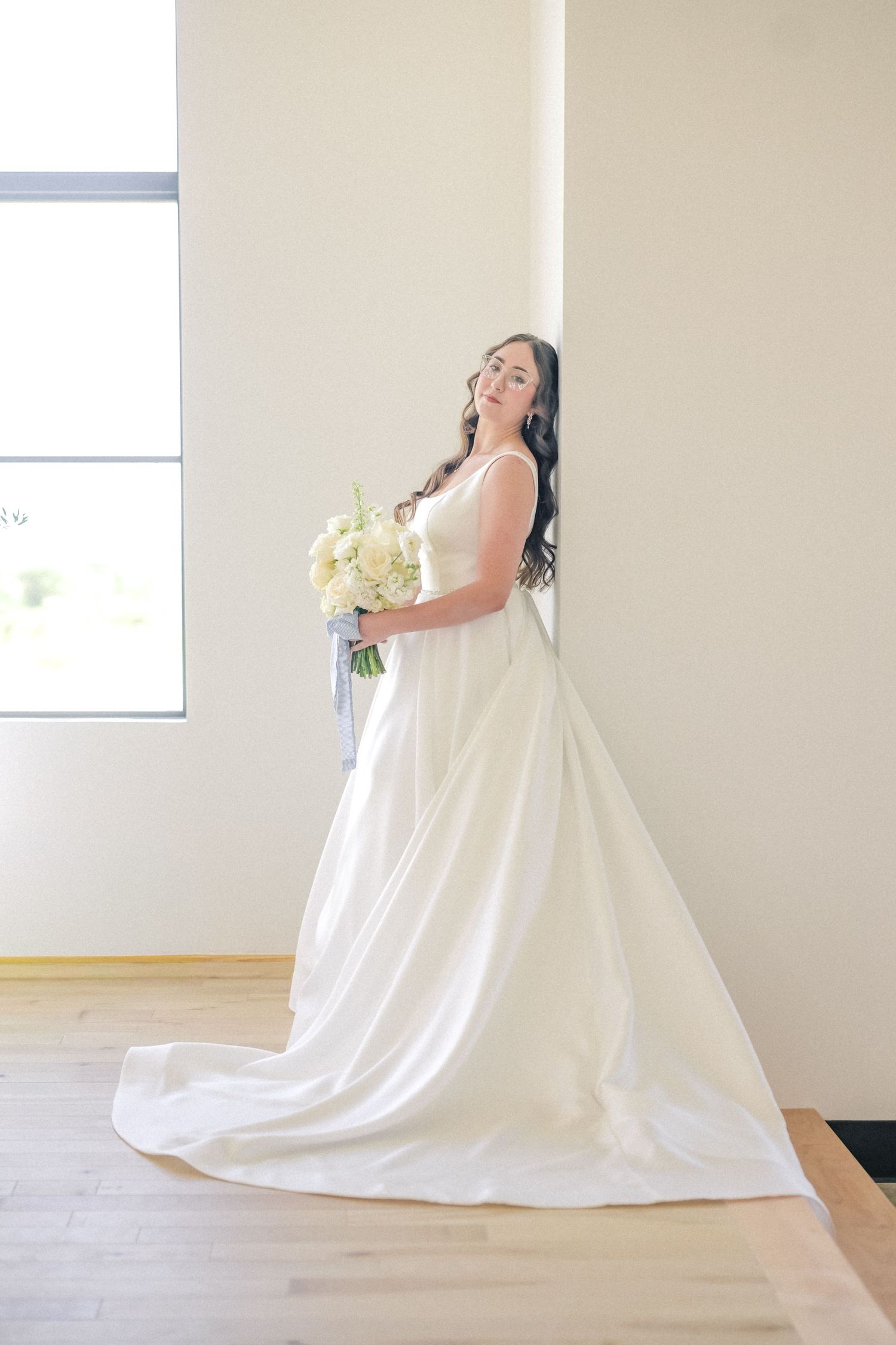 Elegant bride in flowing white gown holding white floral bouquet leaning against wall in bright modern space.