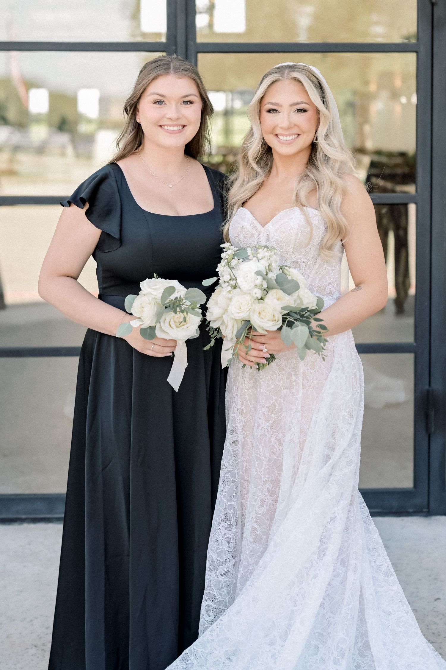 Bride in lace gown and bridesmaid in black dress hold white rose bouquets outside wedding venue.