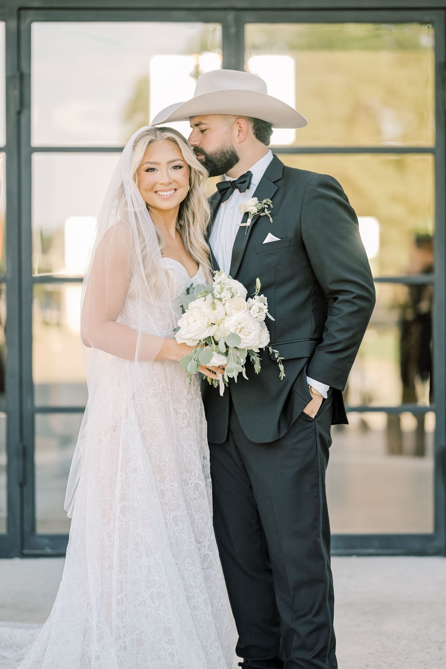 Bride in lace gown and groom in black suit with cowboy hat pose with white bouquet.
