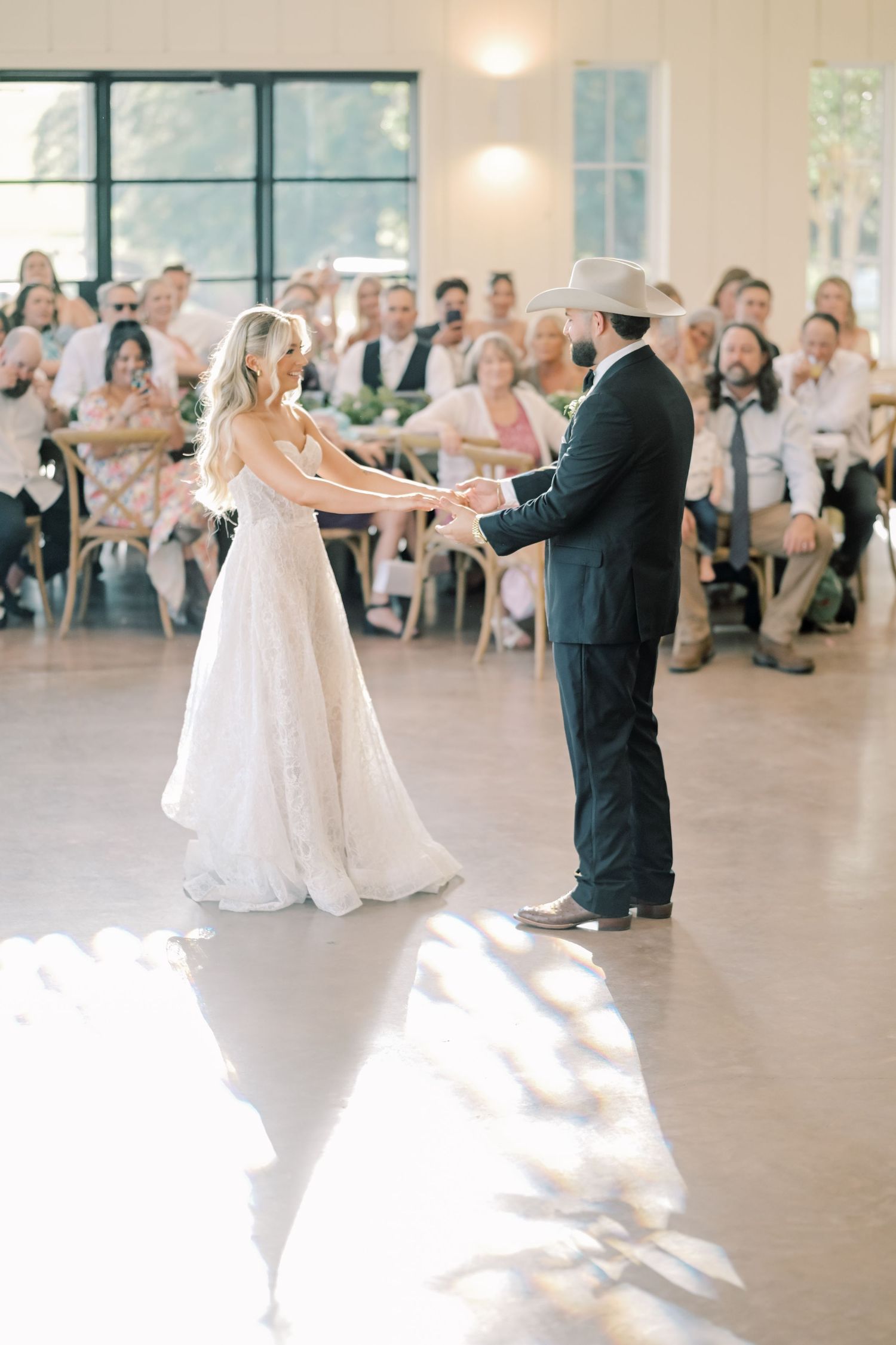 Bride and groom share their first dance in a sunlit wedding reception hall with guests watching.