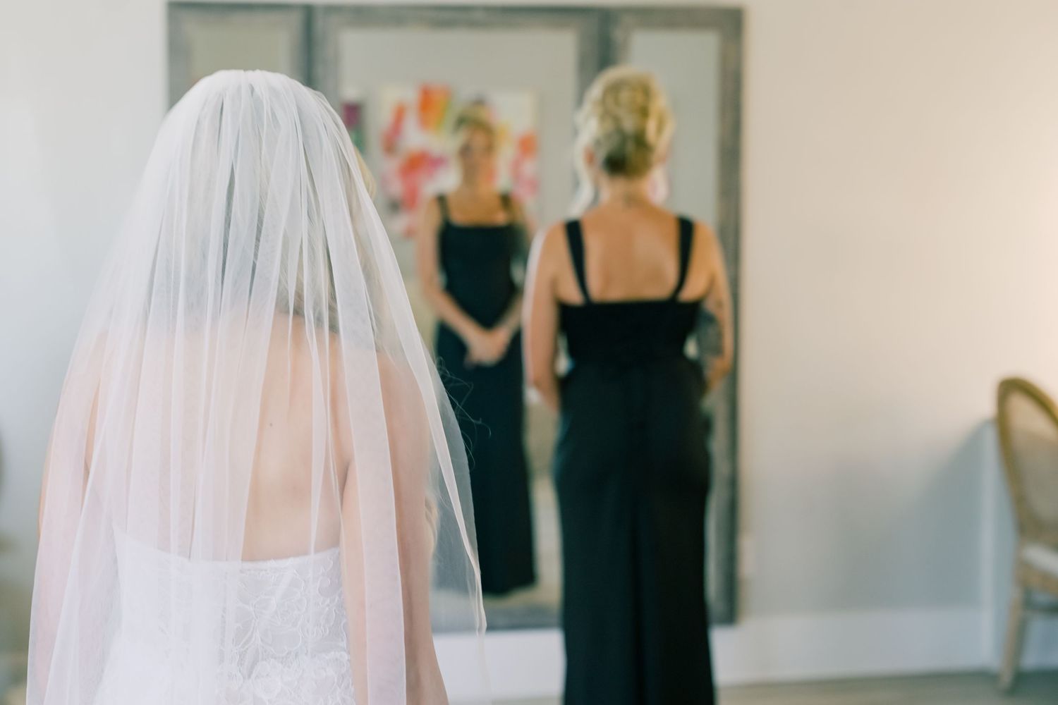 Bride in white gown and veil stands before mirror with bridesmaid in black dress.