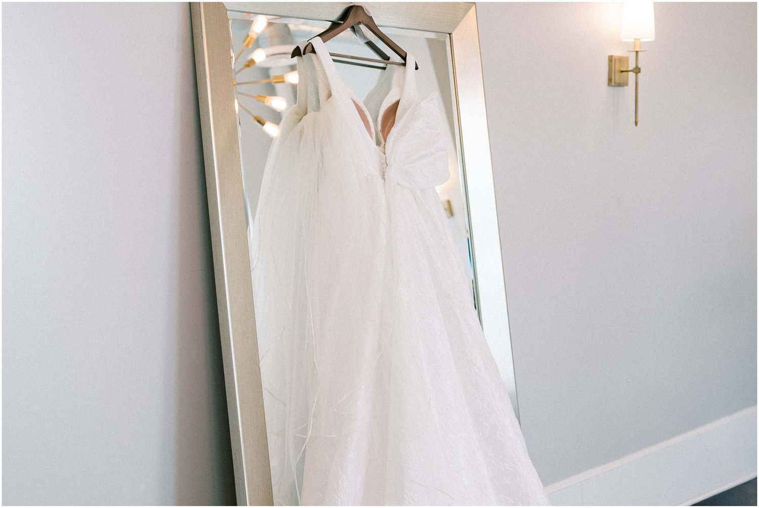White wedding gown displayed on hanger against neutral wall in morning light.