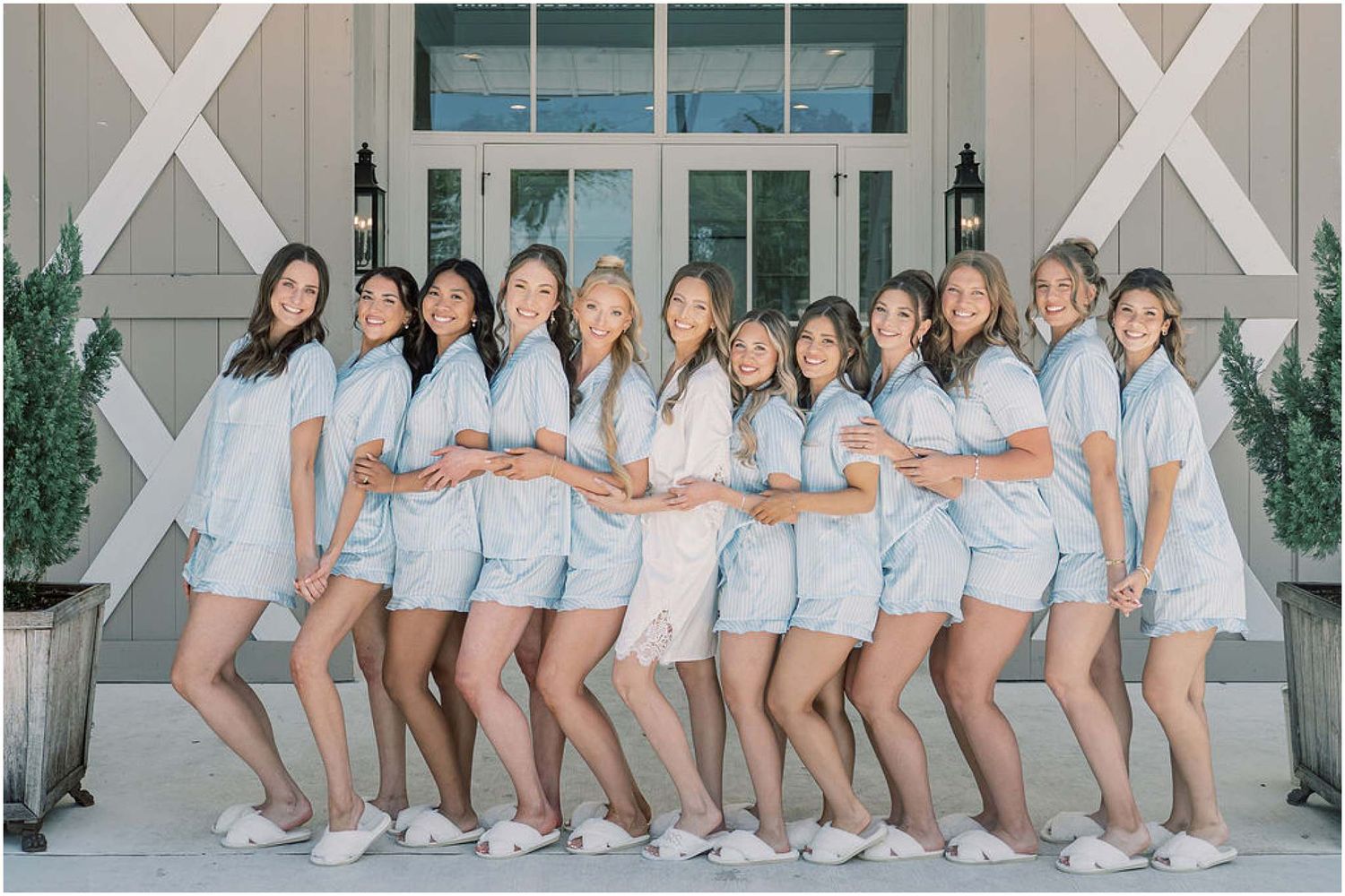 Group of bridesmaids posing together in matching light blue pajama sets and white slippers outside a venue.