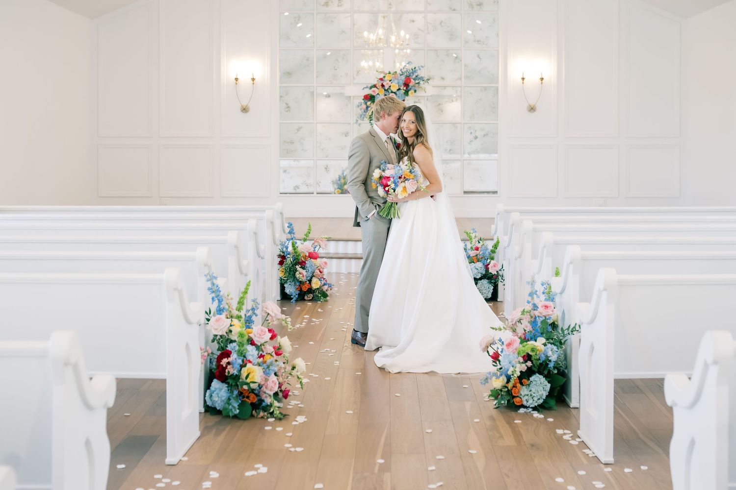 Bride and groom embrace in a bright white chapel aisle adorned with colorful wildflower arrangements on wooden pews.