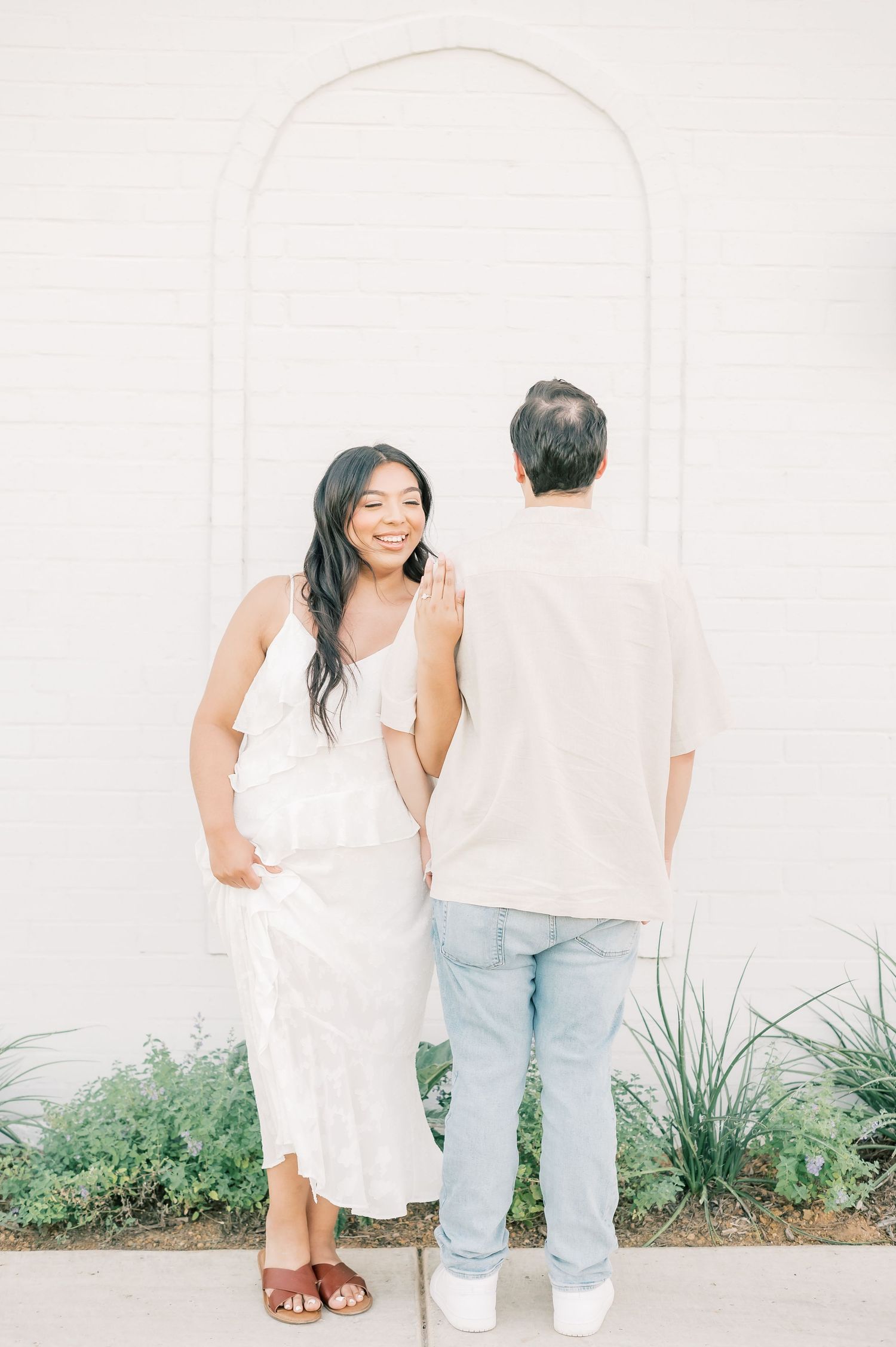 A couple in white and light blue outfits poses playfully against a white wall with green plants at the base.
