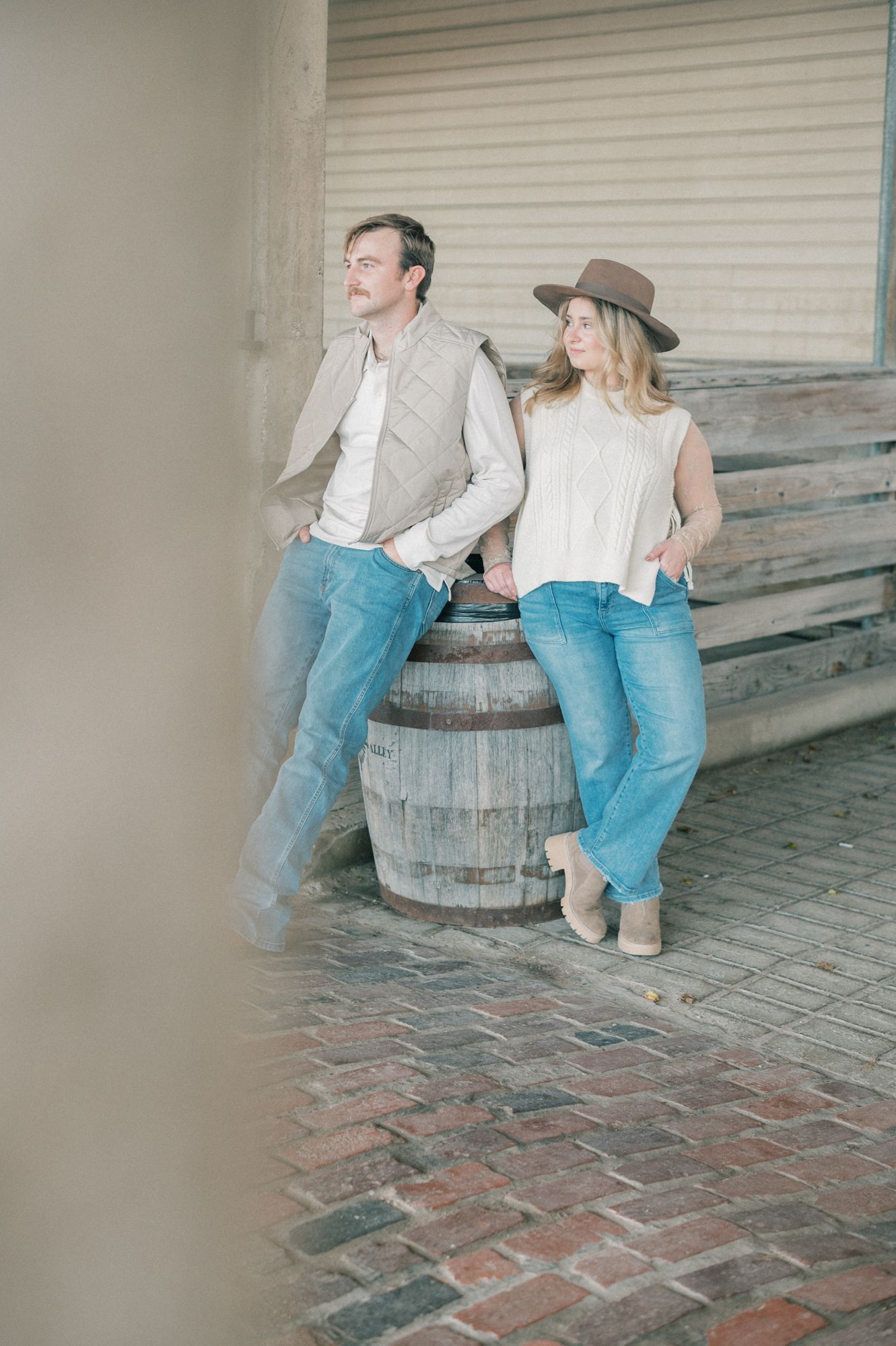 Couple in coordinated outfits leaning against a vintage barrel in an industrial setting.