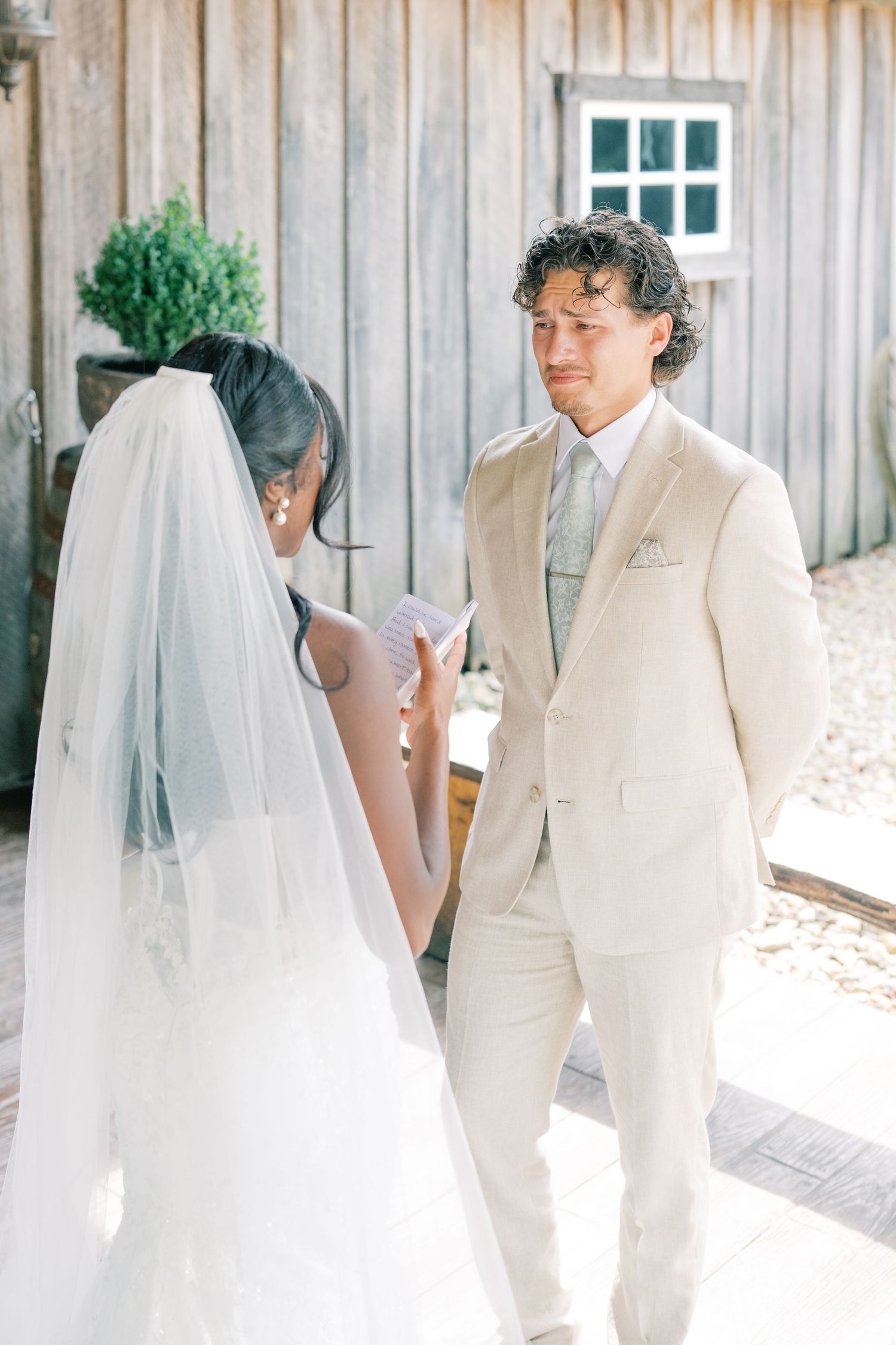 Groom in beige suit listens emotionally as bride reads wedding vows outdoors near rustic barn.