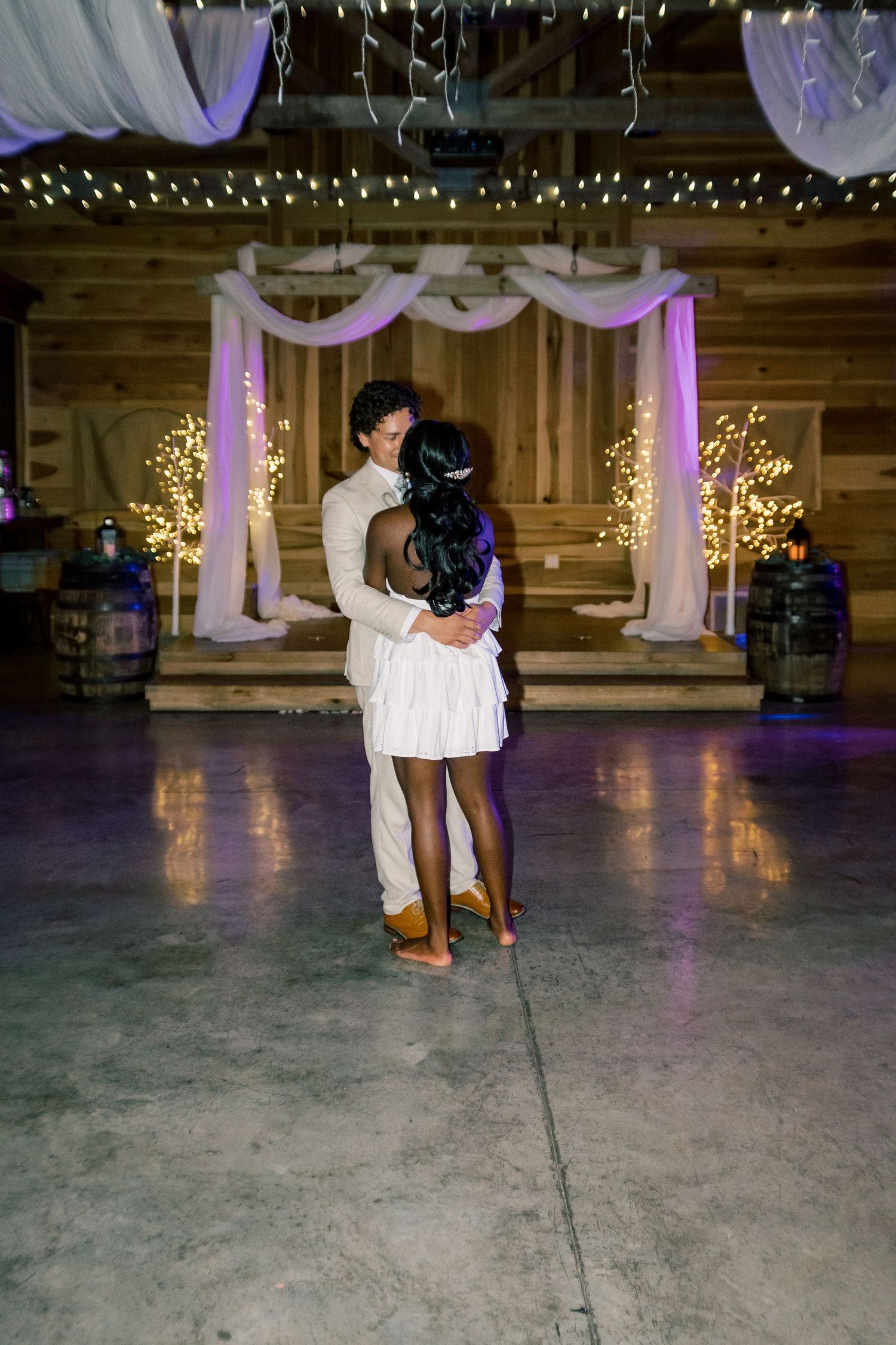 Couple shares romantic first dance in dimly lit barn reception venue with fairy lights and white draping.