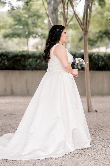 Side profile of bride in white satin A-line wedding dress holding bouquet among trees on gravel path.