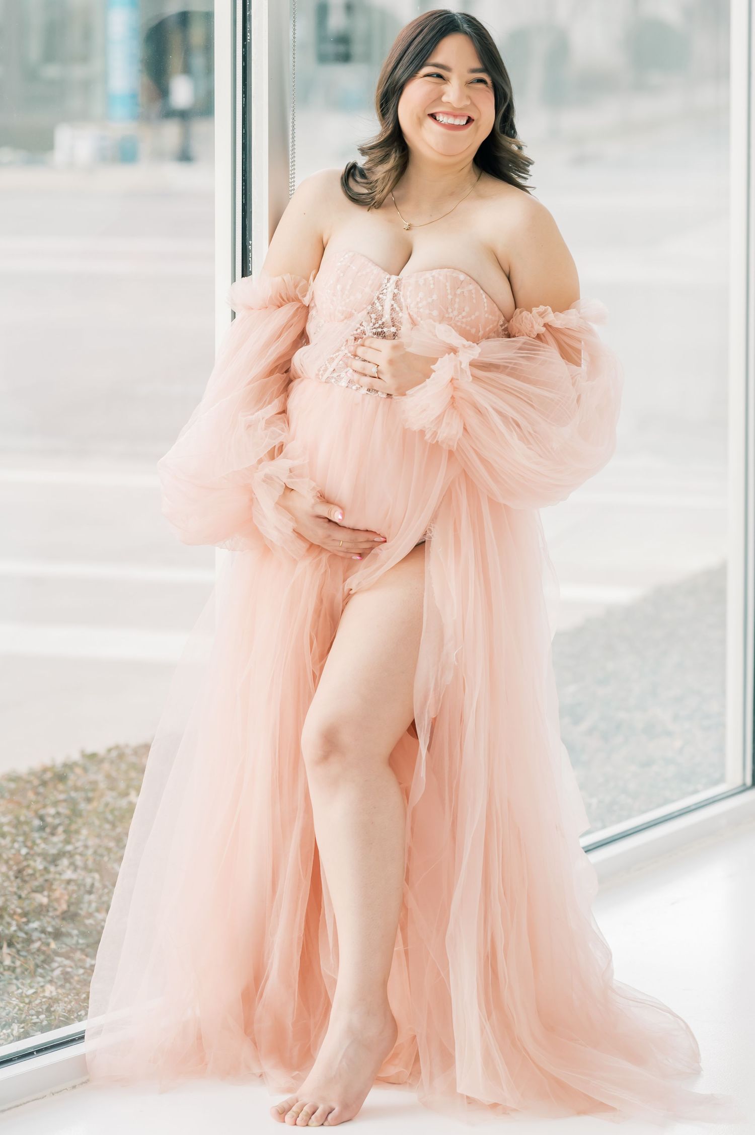 Smiling pregnant woman in a blush pink tulle maternity gown posing near a large window in a bright studio.