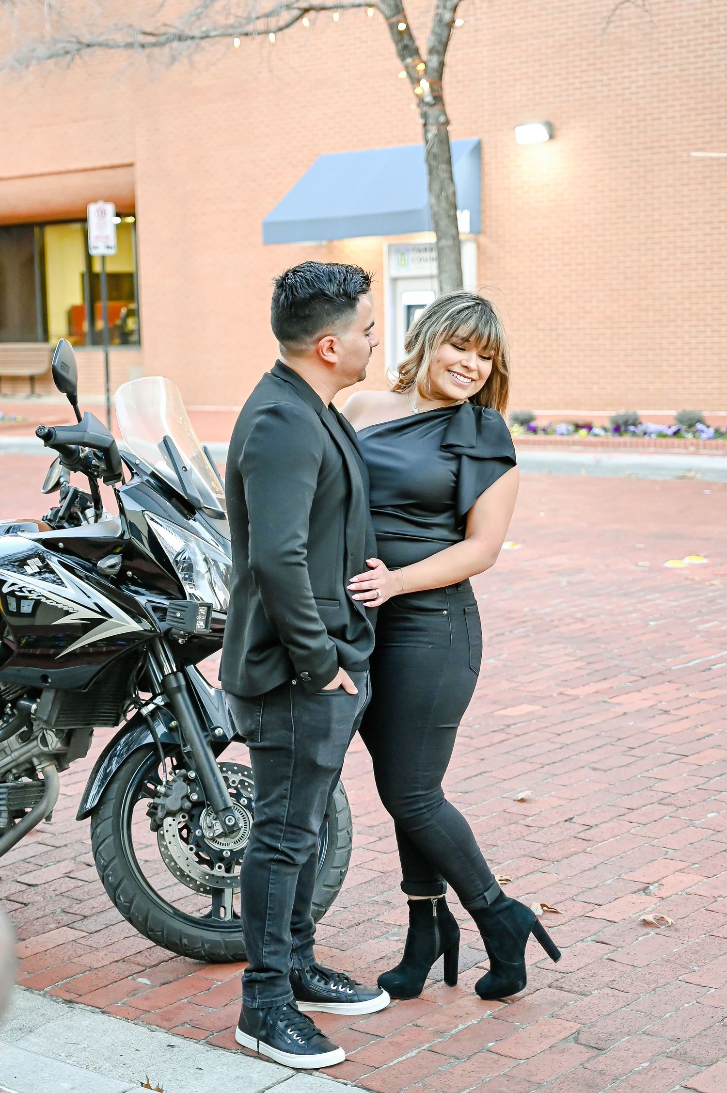 Couple in matching black outfits posing together near a motorcycle on a brick sidewalk.