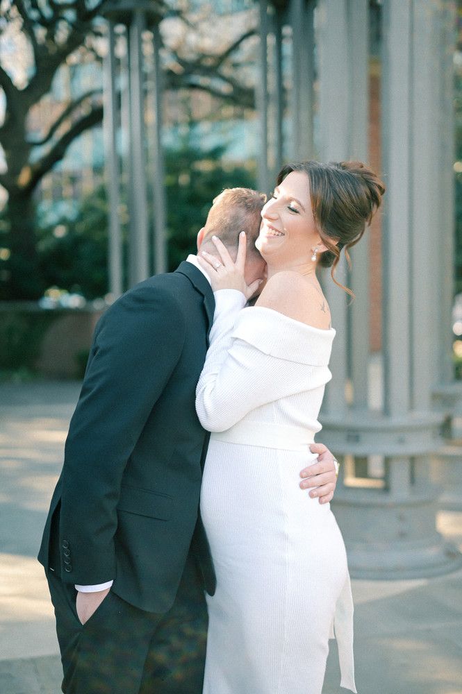 Groom kissing brides neck as she smiles in elegant white off-shoulder wedding gown at outdoor venue.