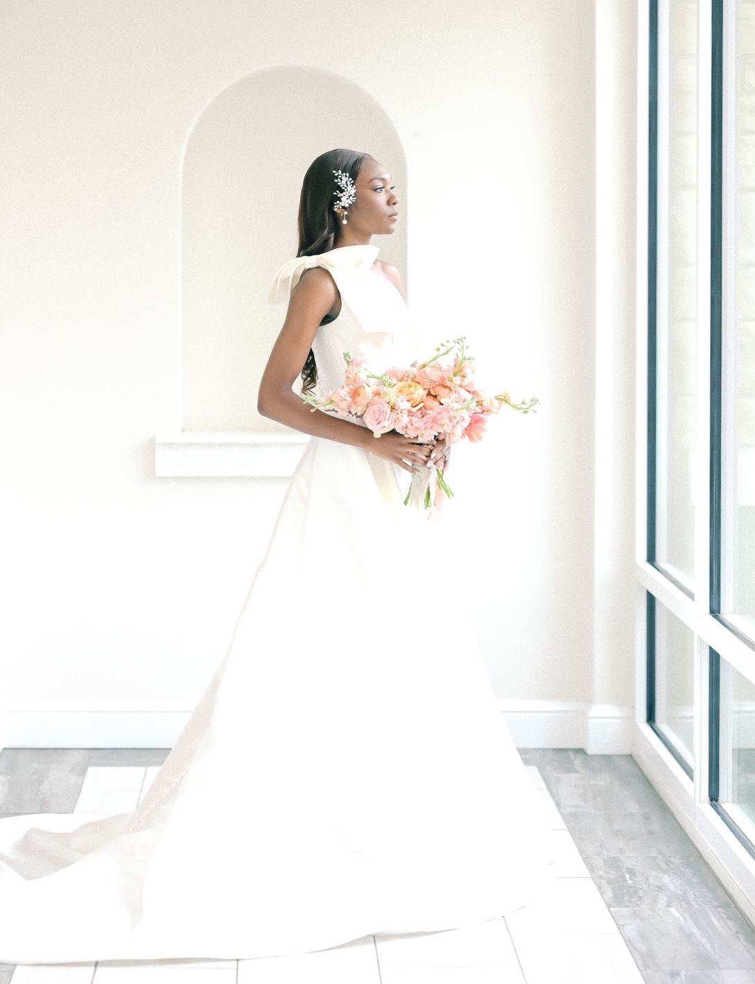 Elegant Black bride in white A-line wedding gown holding pink floral bouquet standing by arched window in bright room.