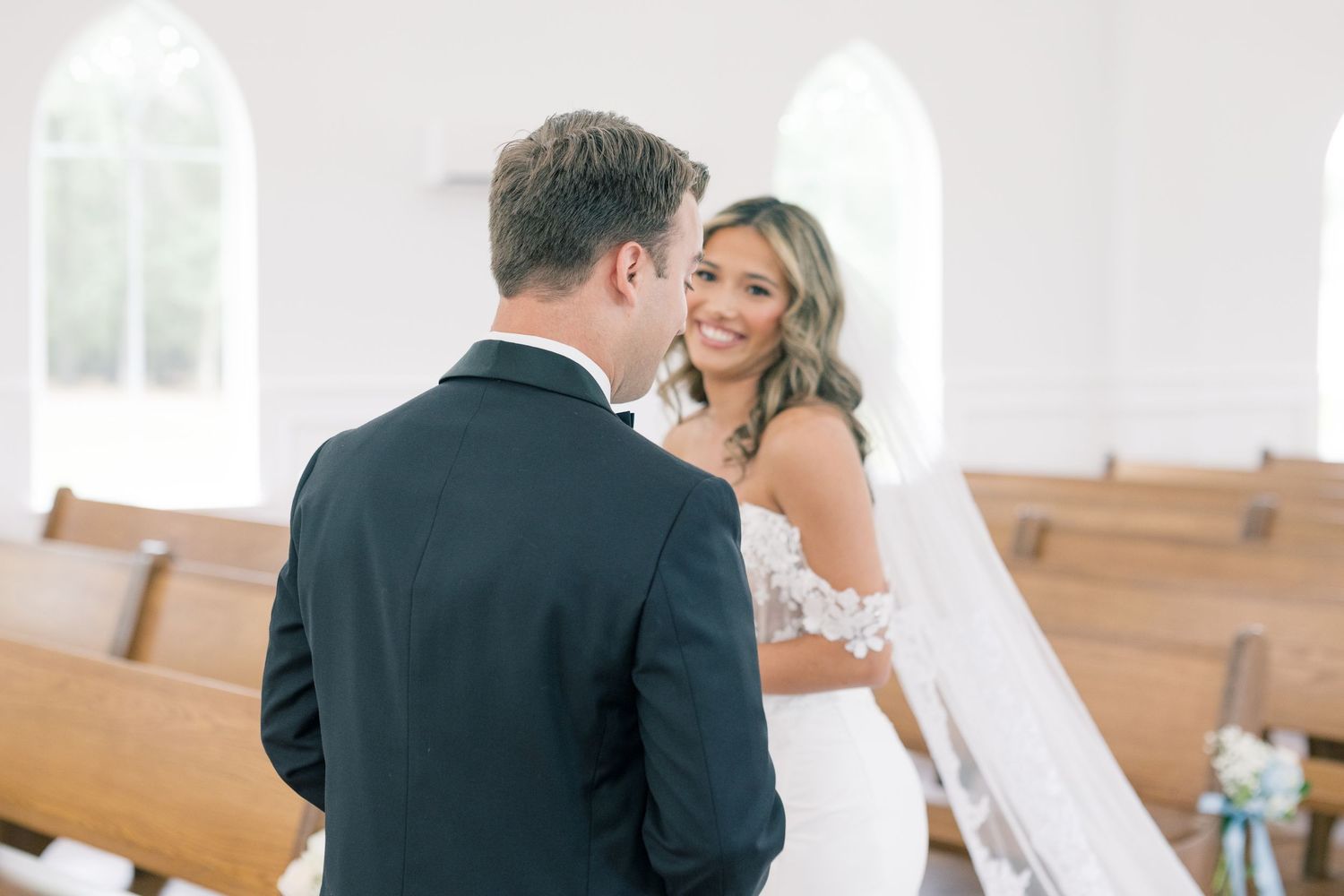 Bride in lace off-shoulder gown smiling at groom inside a white chapel with wooden pews.