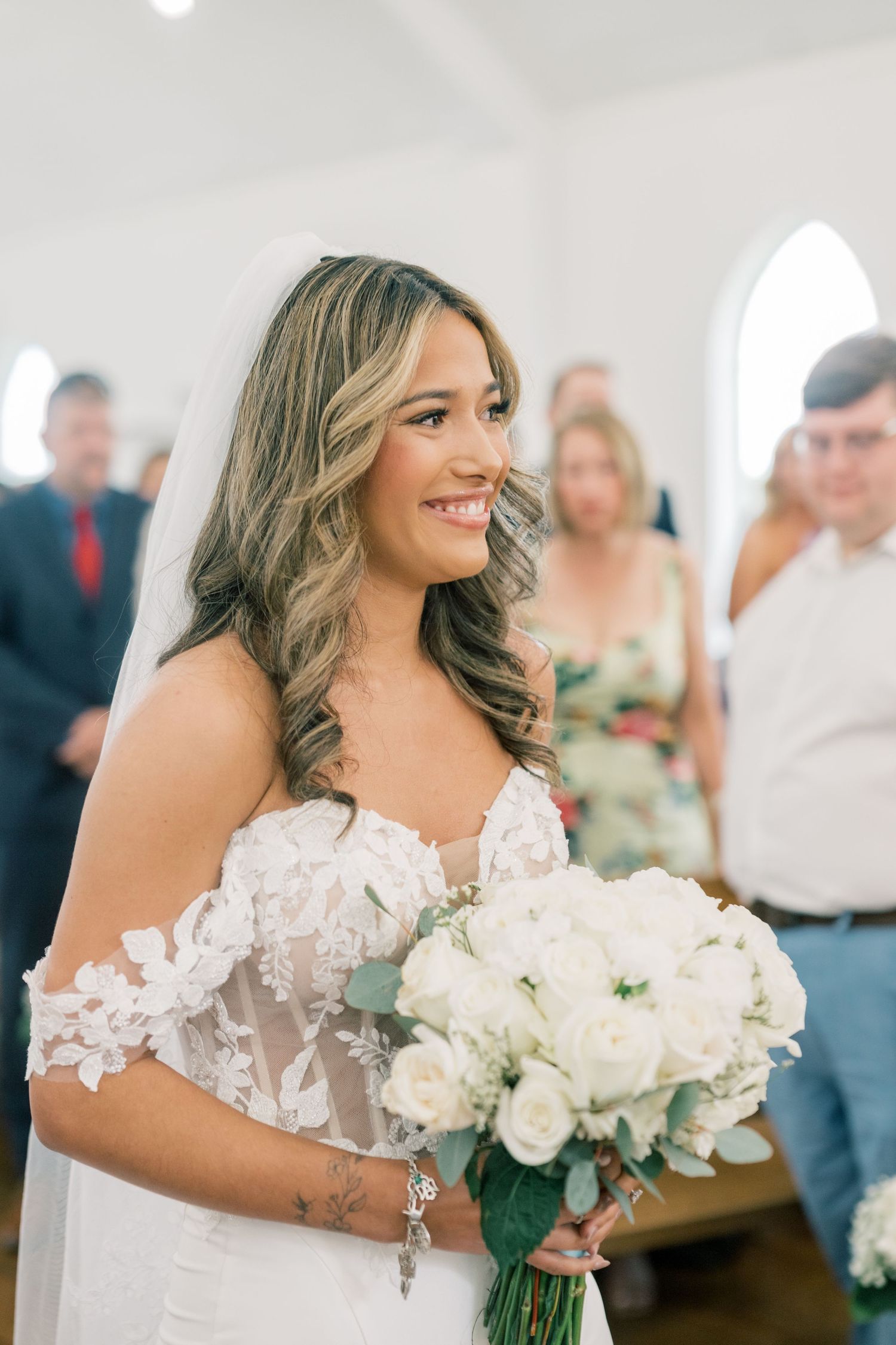 Smiling bride in lace off-shoulder wedding dress holds white rose bouquet while walking down the aisle.