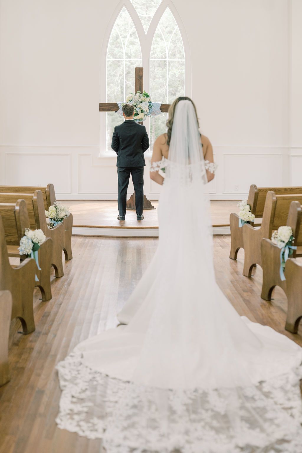 Bride walking down the aisle toward groom in a white chapel with wooden pews and cross.